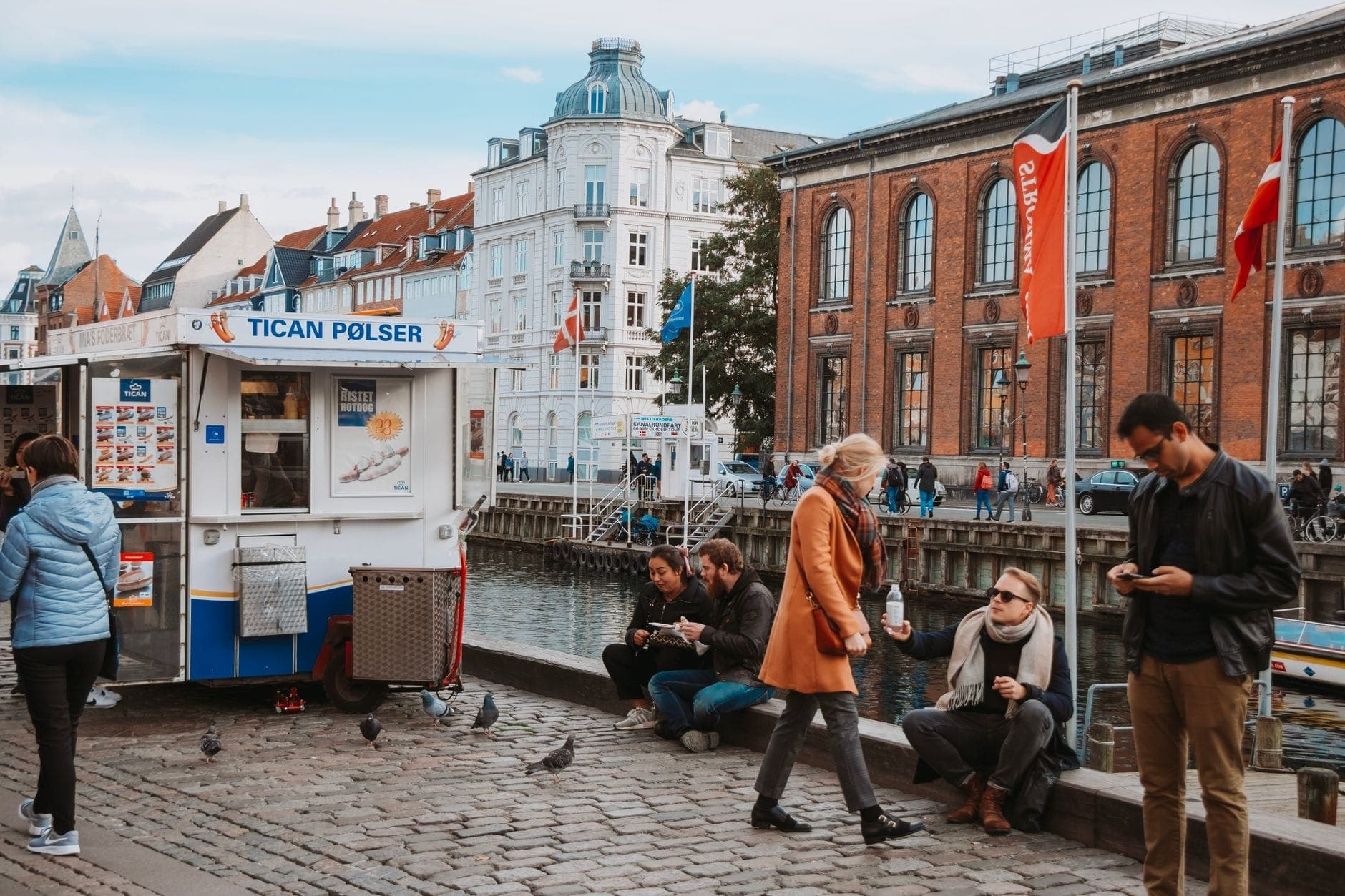 Pessoas relaxando à beira de um canal em Copenhague com uma barraca de comida de rua, bandeiras da cidade e edifícios históricos sob a luz do dia.