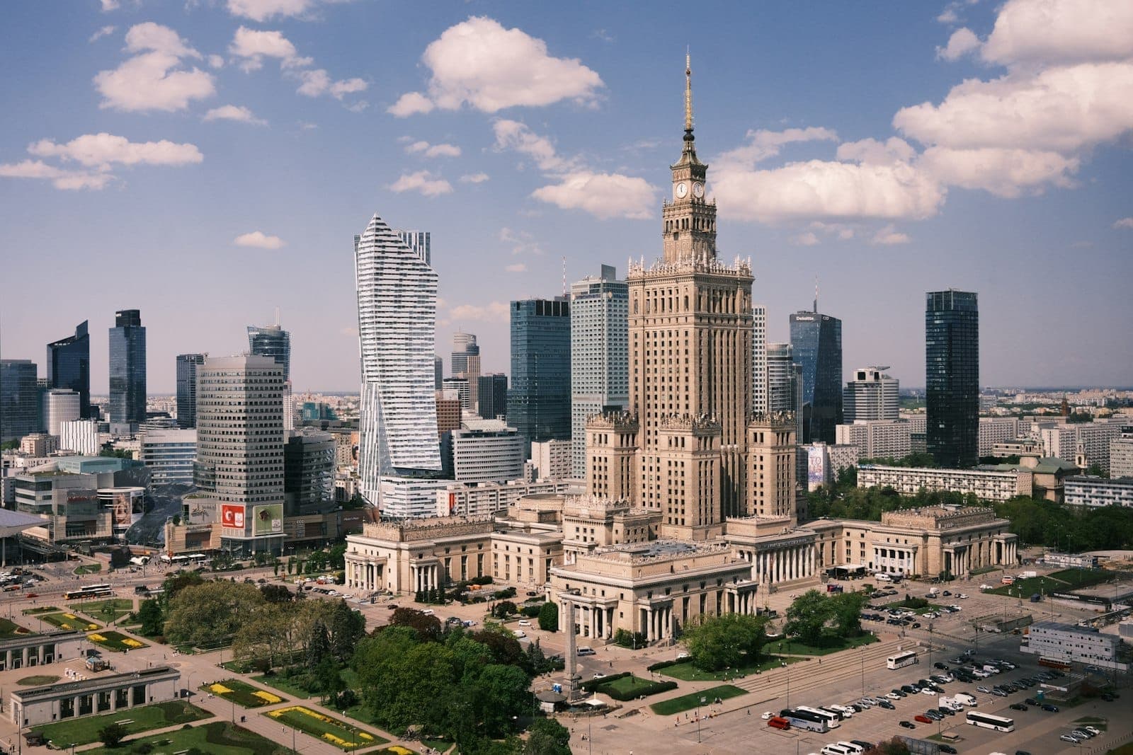 Dzienny widok na panoramę centrum Warszawy z Pałacem Kultury i Nauki otoczonym nowoczesnymi wieżowcami i terenami zielonymi.