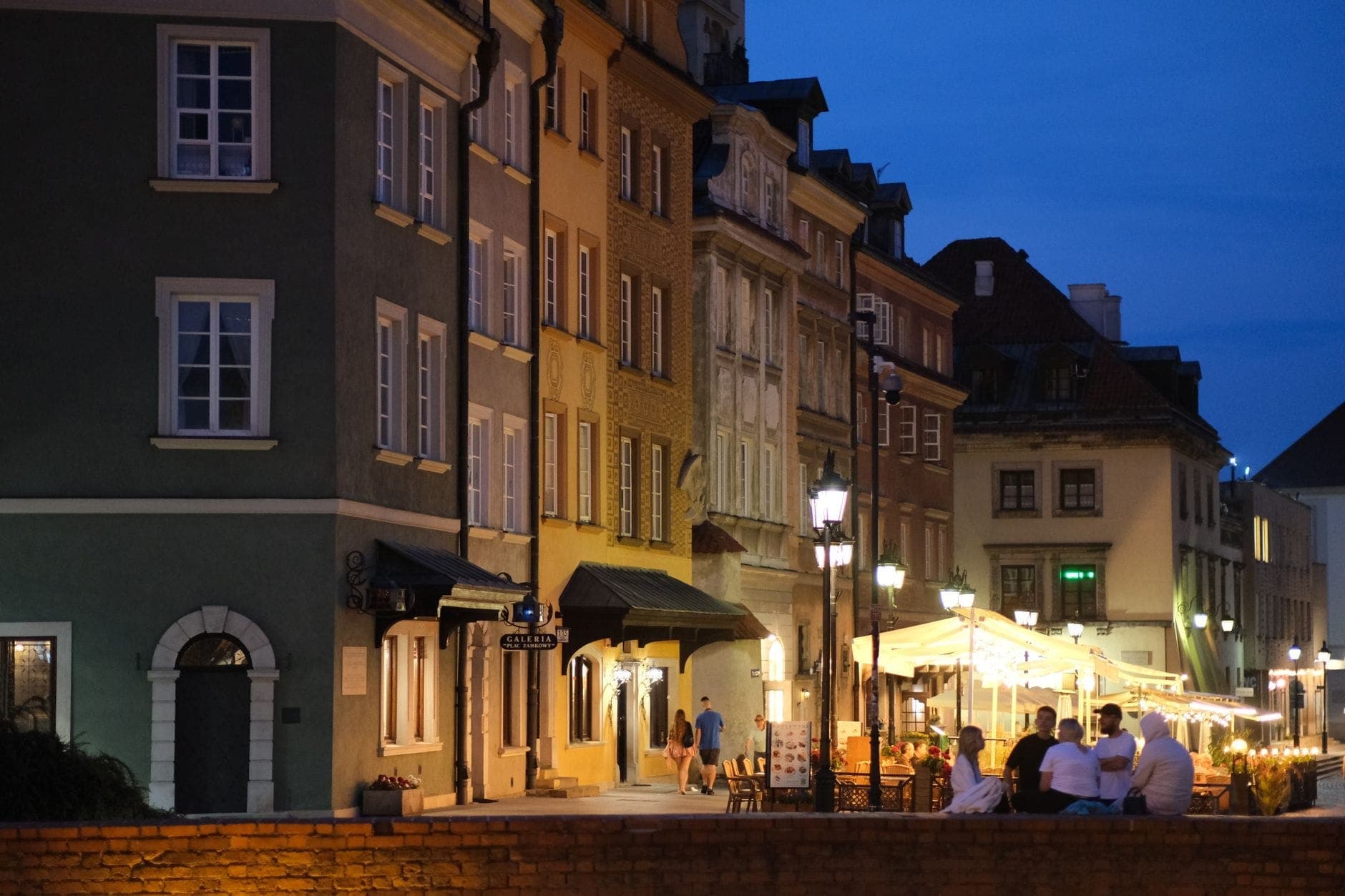 Wieczorna scena uliczna w Warszawie z kolorowymi historycznymi budynkami, restauracjami na świeżym powietrzu, ciepłym oświetleniem i ludźmi jedzącymi kolację pod markizami.