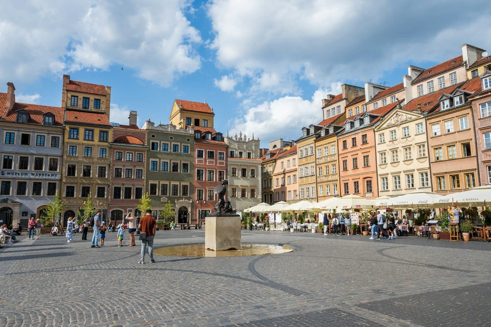 Weiter Blick auf den Warschauer Altstadtmarkt an einem sonnigen Tag mit bunten historischen Gebäuden, der Sigismundsäule, Spaziergängern und blauem Himmel mit Wolken.