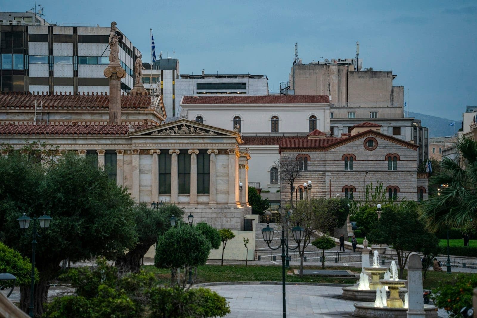 Neoklassizistische Gebäude und Statue auf einem Platz in einem historischen Viertel in Athen, umgeben von Stadtarchitektur und Grün in der Abenddämmerung.