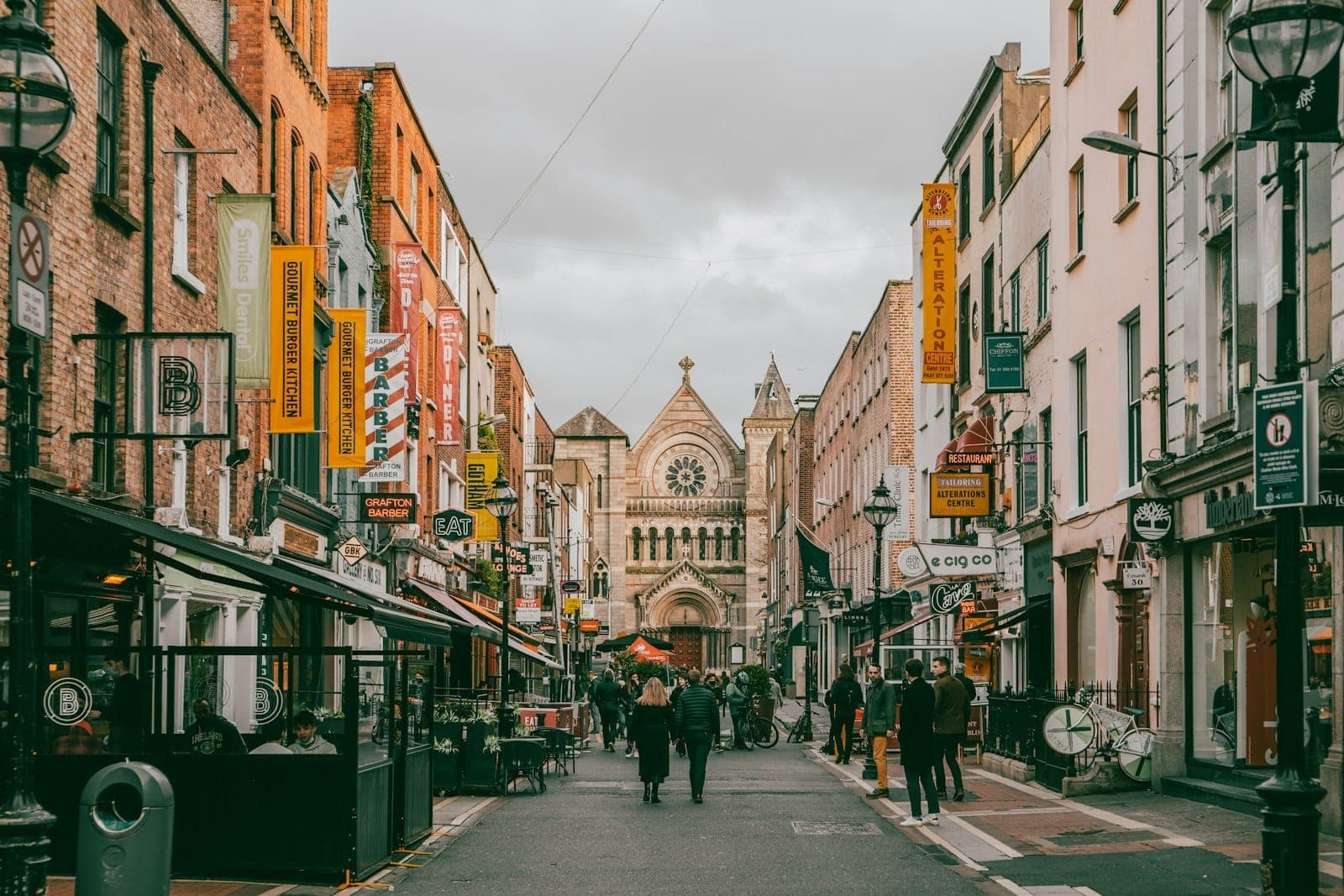 Belebte Fußgängerzone in Dublin mit Menschen vor Cafés und Geschäften, bunten Hausfassaden und einer historischen Kirche am Ende der Straße.
