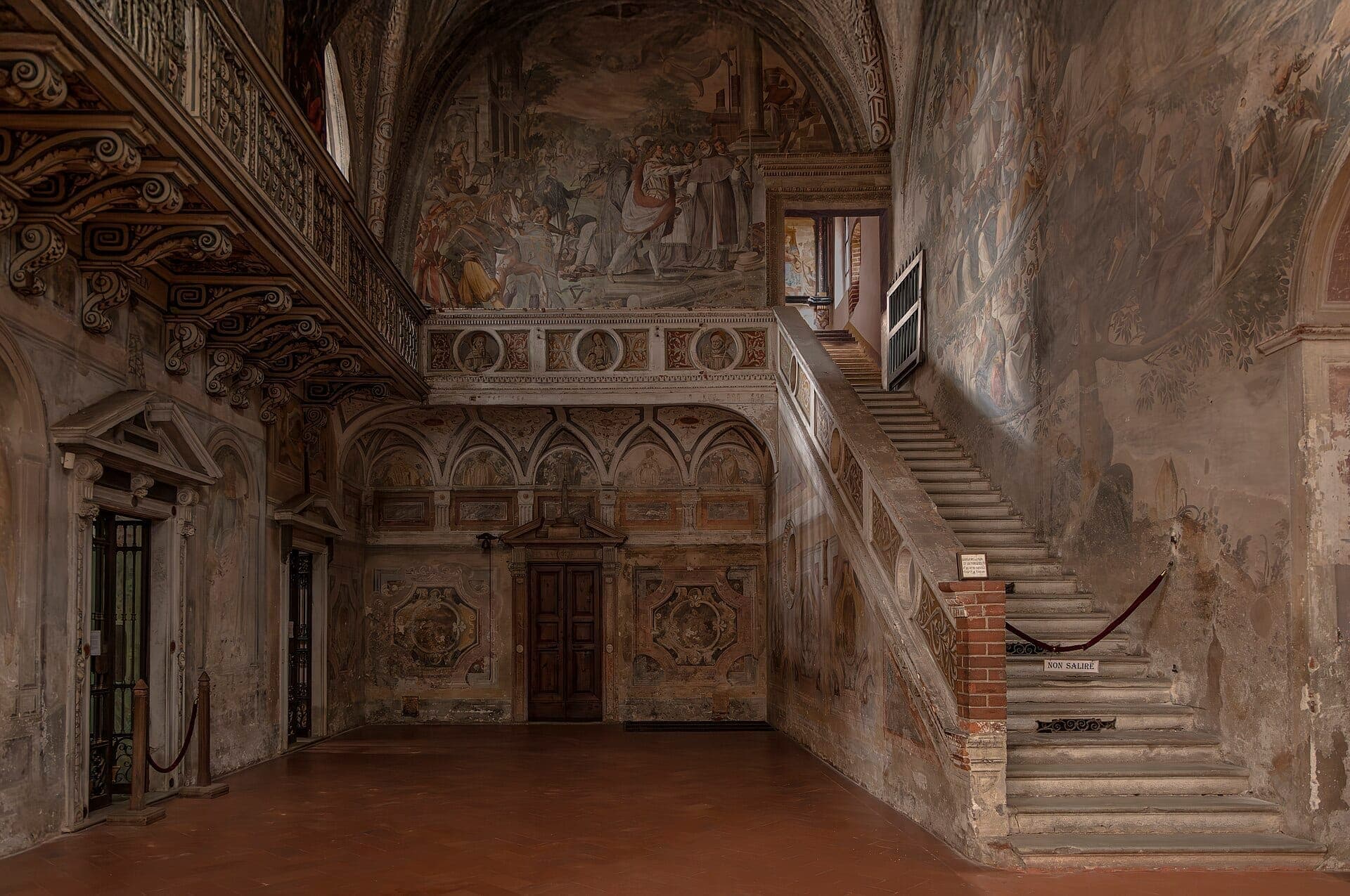 Ornate frescoed interior and stone staircase of Abbazia di Chiaravalle, featuring detailed wall paintings, arched ceilings, and warm ambient lighting.