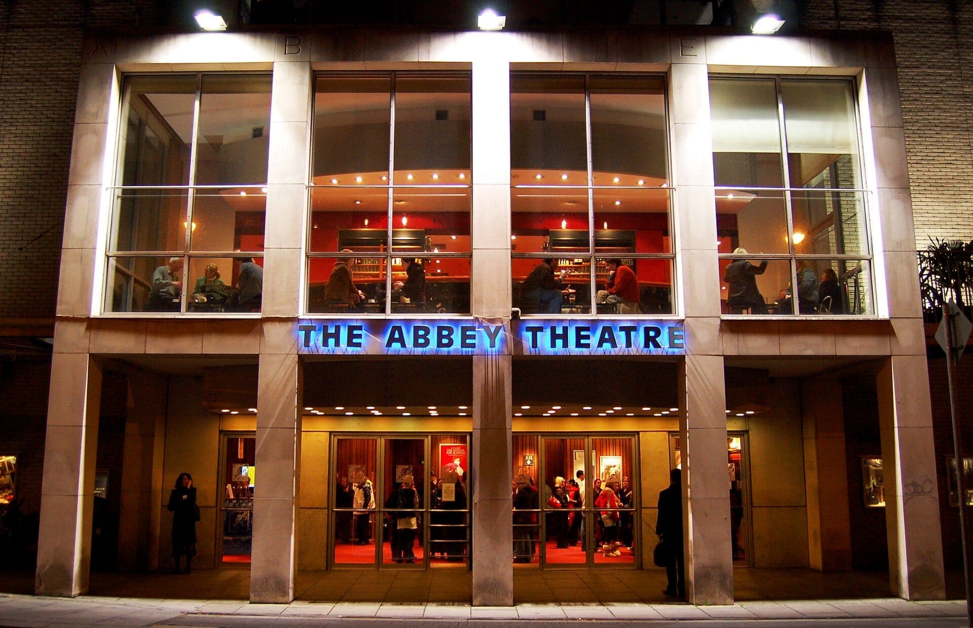 L'ingresso principale dell'Abbey Theatre a Dublino di notte, con insegne illuminate, grandi vetrate e persone visibili all'interno.