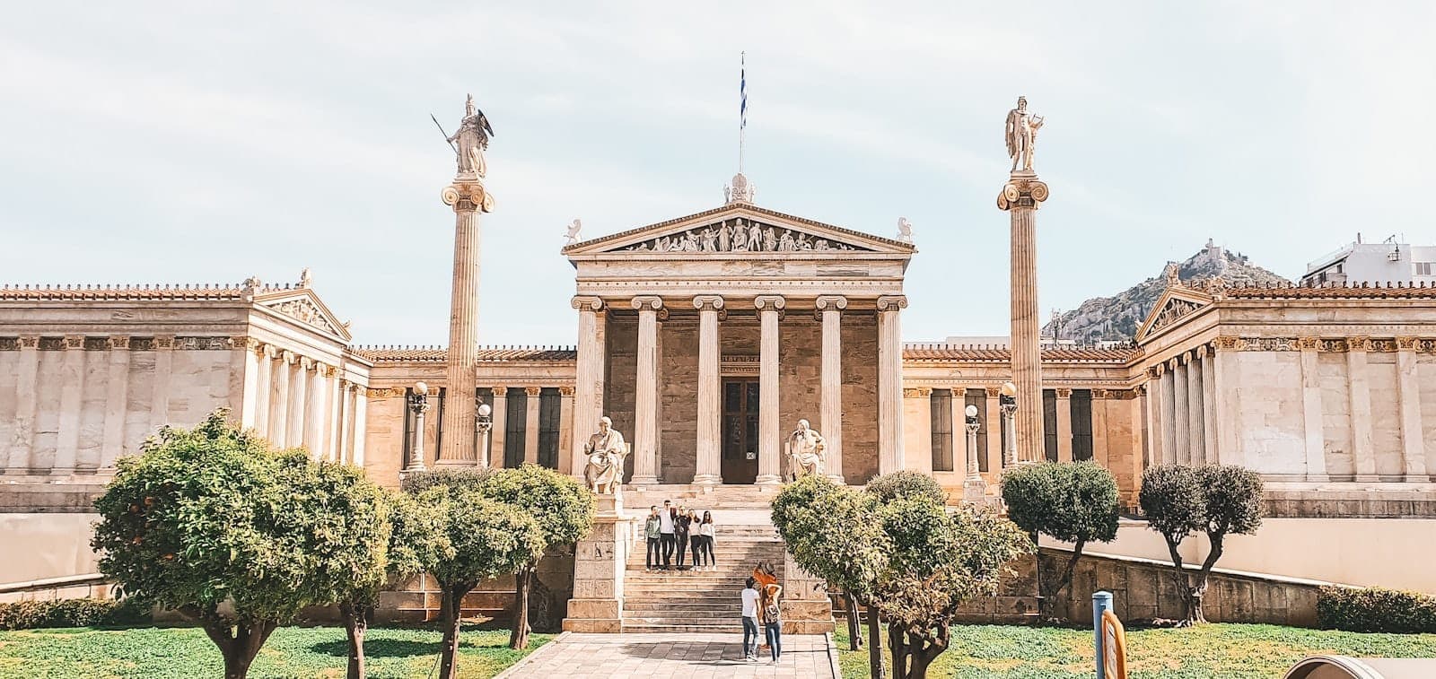 Wide view of the Athenian Academy with neoclassical columns, statues, and lush greenery, showing the stunning architecture on a bright, clear day.