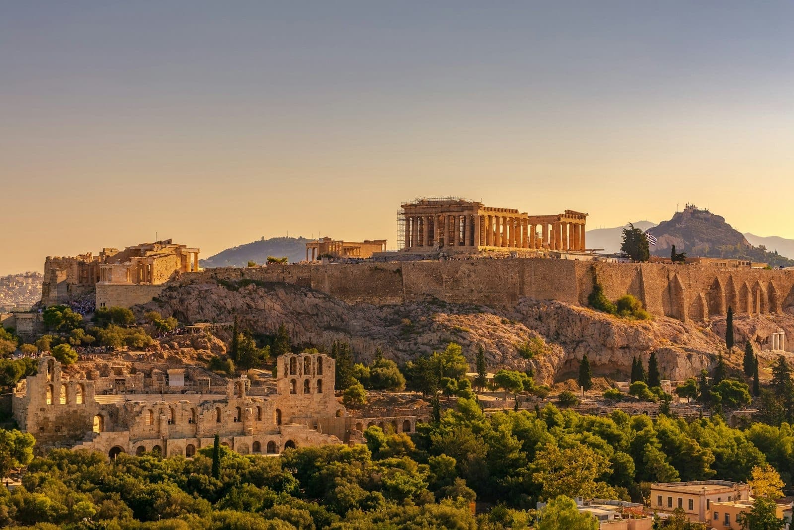 Blick auf die Akropolis von Athen bei Sonnenuntergang, mit dem Parthenon und umliegenden antiken Ruinen über üppigem Grün und der Stadtlandschaft.