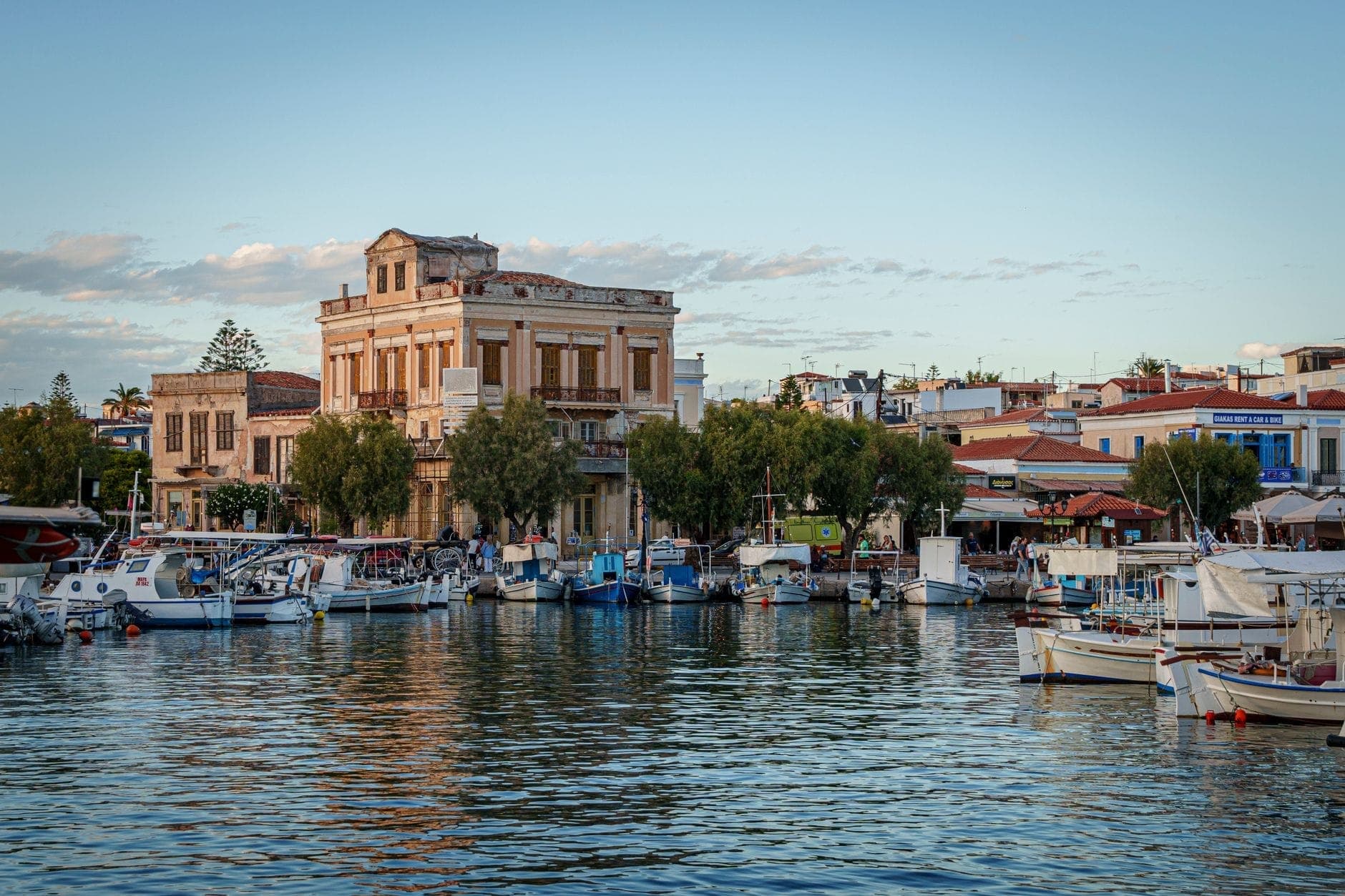 Aegina Island’s charming waterfront with traditional buildings, small boats moored at the lively port, and clear reflections on the water under a bright blue sky.