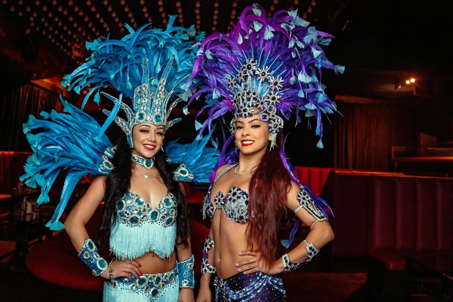 Two performers in elaborate feathered costumes with sparkling details stand smiling inside a cabaret show venue with dark red seating.