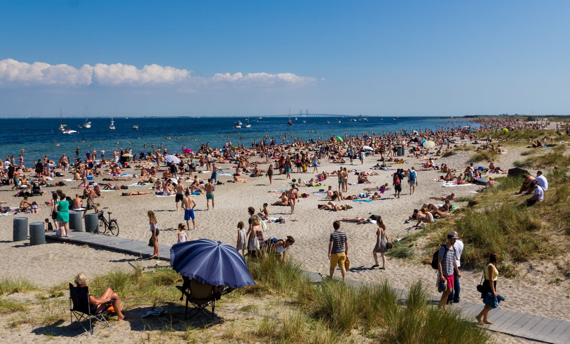 Menschenmassen genießen die Sonne am Sandstrand des Amager Strandpark, mit Menschen beim Sonnenbaden, Spazierengehen und Entspannen nahe dem blauen Meer unter einem klaren Himmel.
