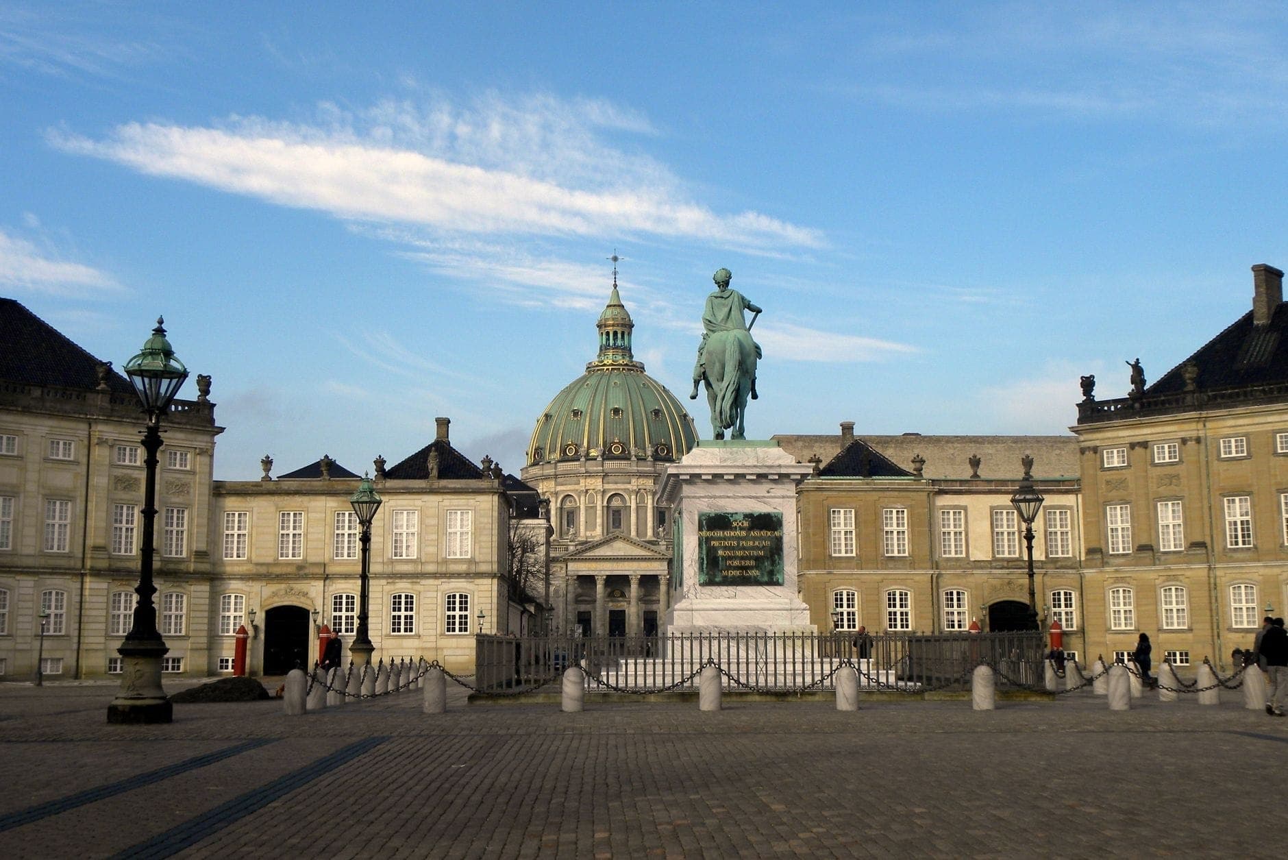 Weitläufige Ansicht des Schlosshofs von Amalienborg mit dem Reiterstandbild in der Mitte und der Kuppel der Frederikskirche im Hintergrund unter einem klaren blauen Himmel.