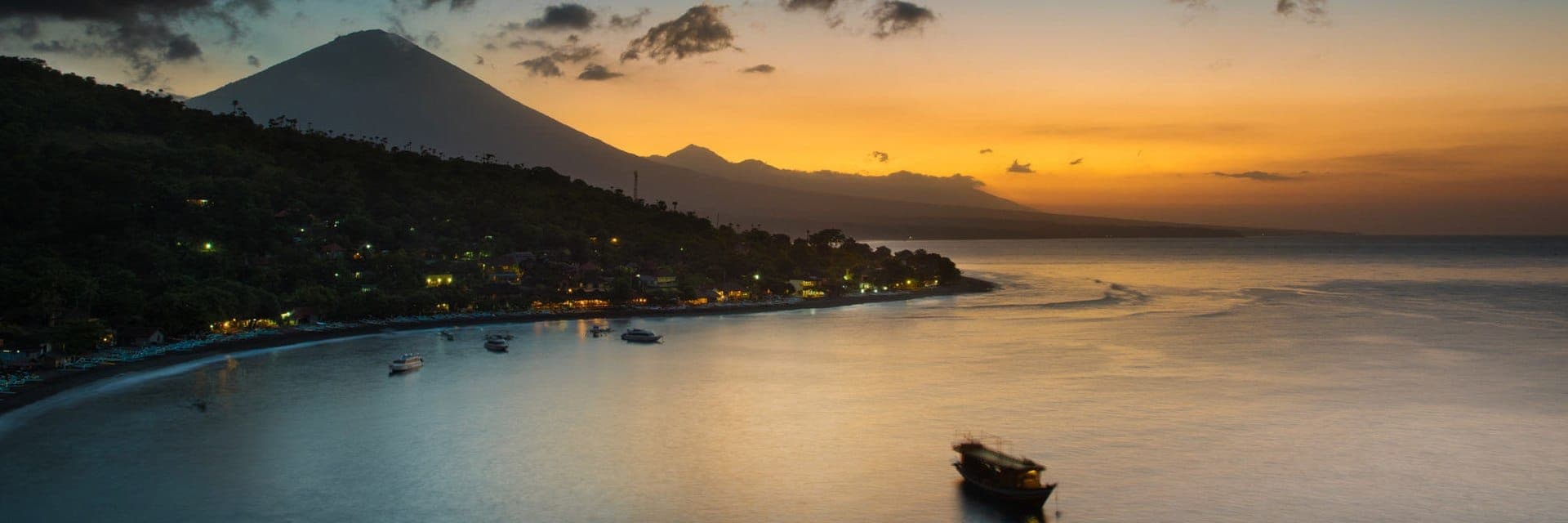 Hoàng hôn yên bình trên Amed, Bali, ánh vàng phủ lên bờ biển sỏi đá, thuyền đánh cá và bóng dáng hùng vĩ của núi Agung.