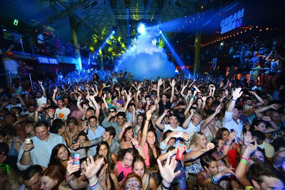 Large crowd dancing under colorful lights and fog inside Amnesia Ibiza nightclub, with raised hands and vibrant party atmosphere.