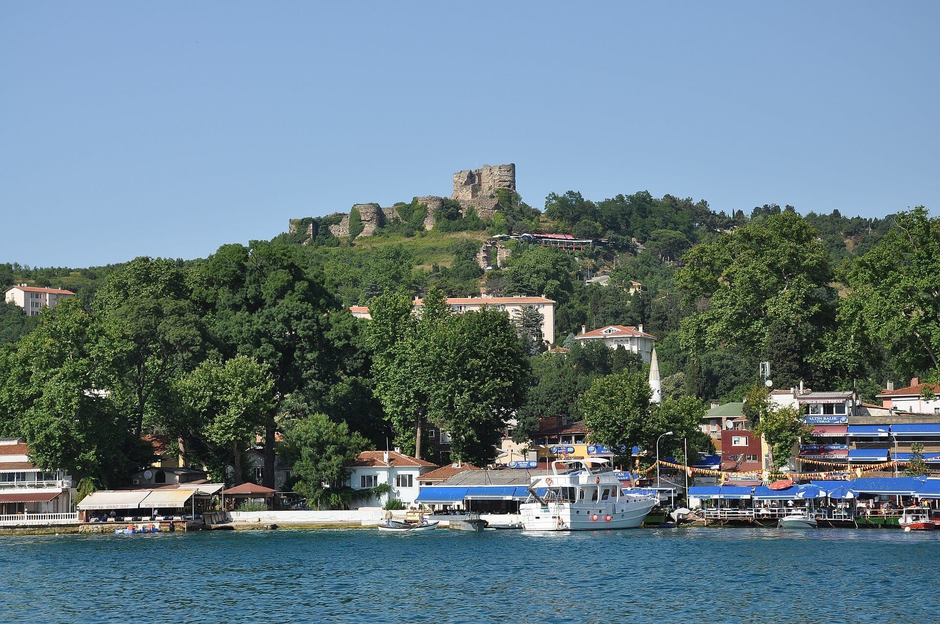 Blick auf das Yoros Castle auf einem grünen Hügel mit Blick auf das bunte Dorf und die Fischerboote von Anadolu Kavağı am Ufer des Bosporus.