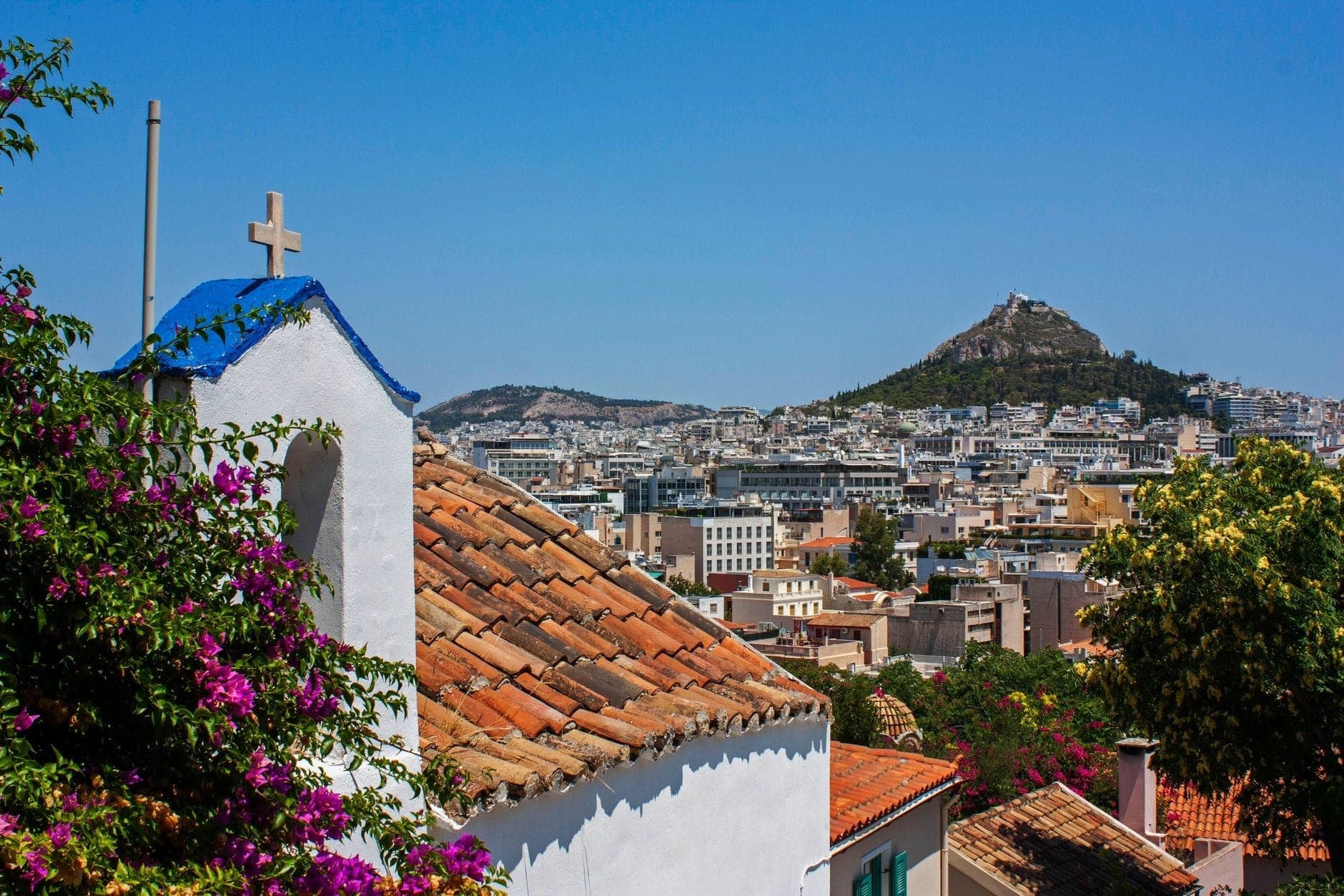 Sunlit view of whitewashed houses with terracotta roofs, a blue-domed chapel, blooming bougainvillea, and Athens cityscape with a hill in the background.