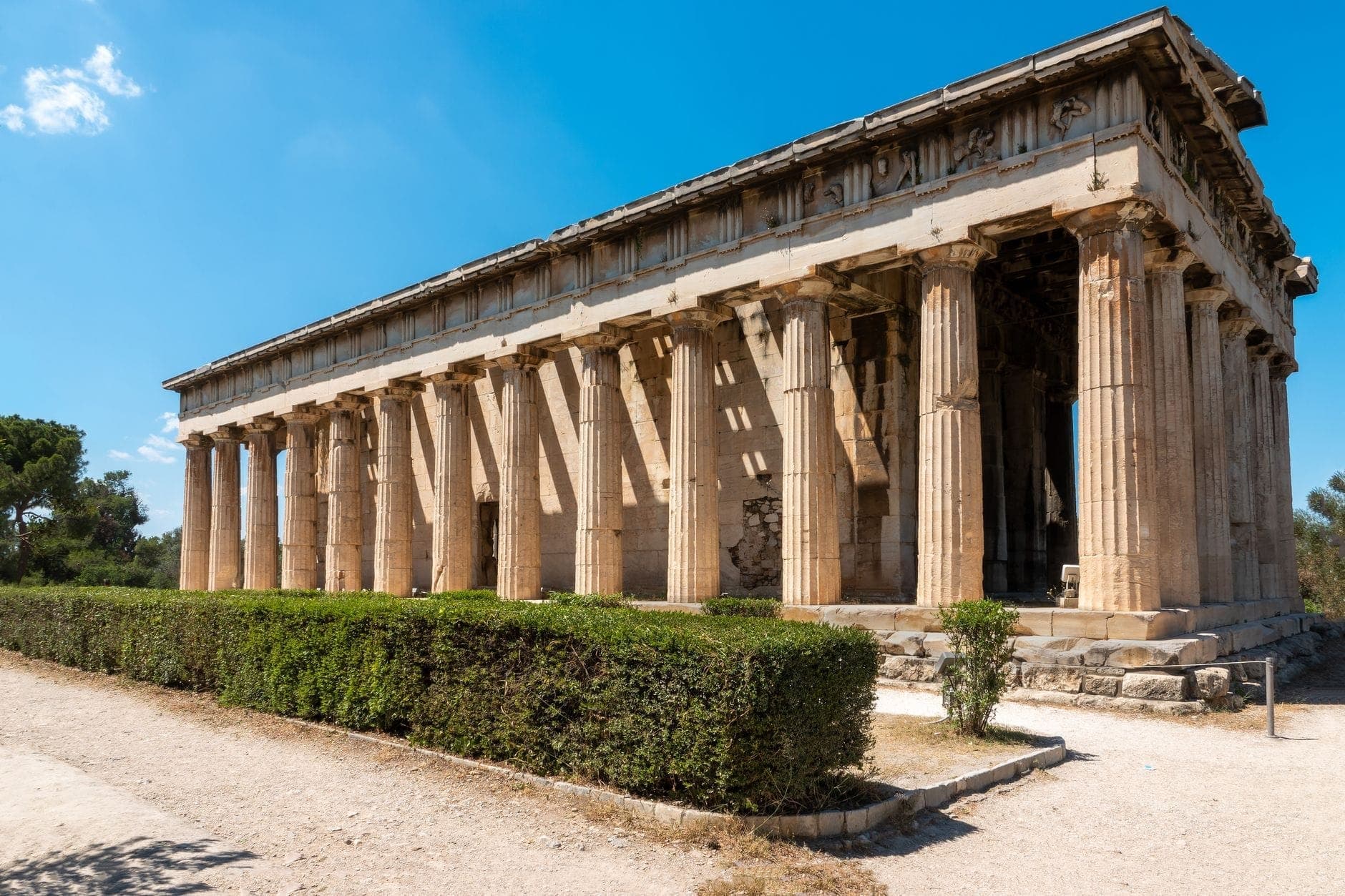 Side view of the well-preserved Temple of Hephaestus at the Ancient Agora of Athens, with blue sky and lush greenery surrounding the ruins.