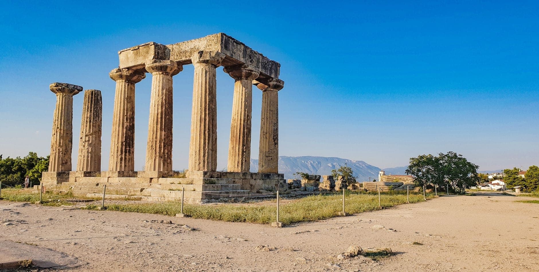 Wide-angle view of the well-preserved ancient Greek temple ruins of Corinth, with seven standing Doric columns under a clear blue sky and distant hills.