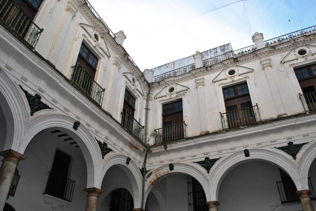 Sân trong của Antiguo Hospital de Mujeres tại Cádiz, với những vòm cung Baroque, cột trụ và mặt tiền trắng cũ kỹ dưới bầu trời xanh.