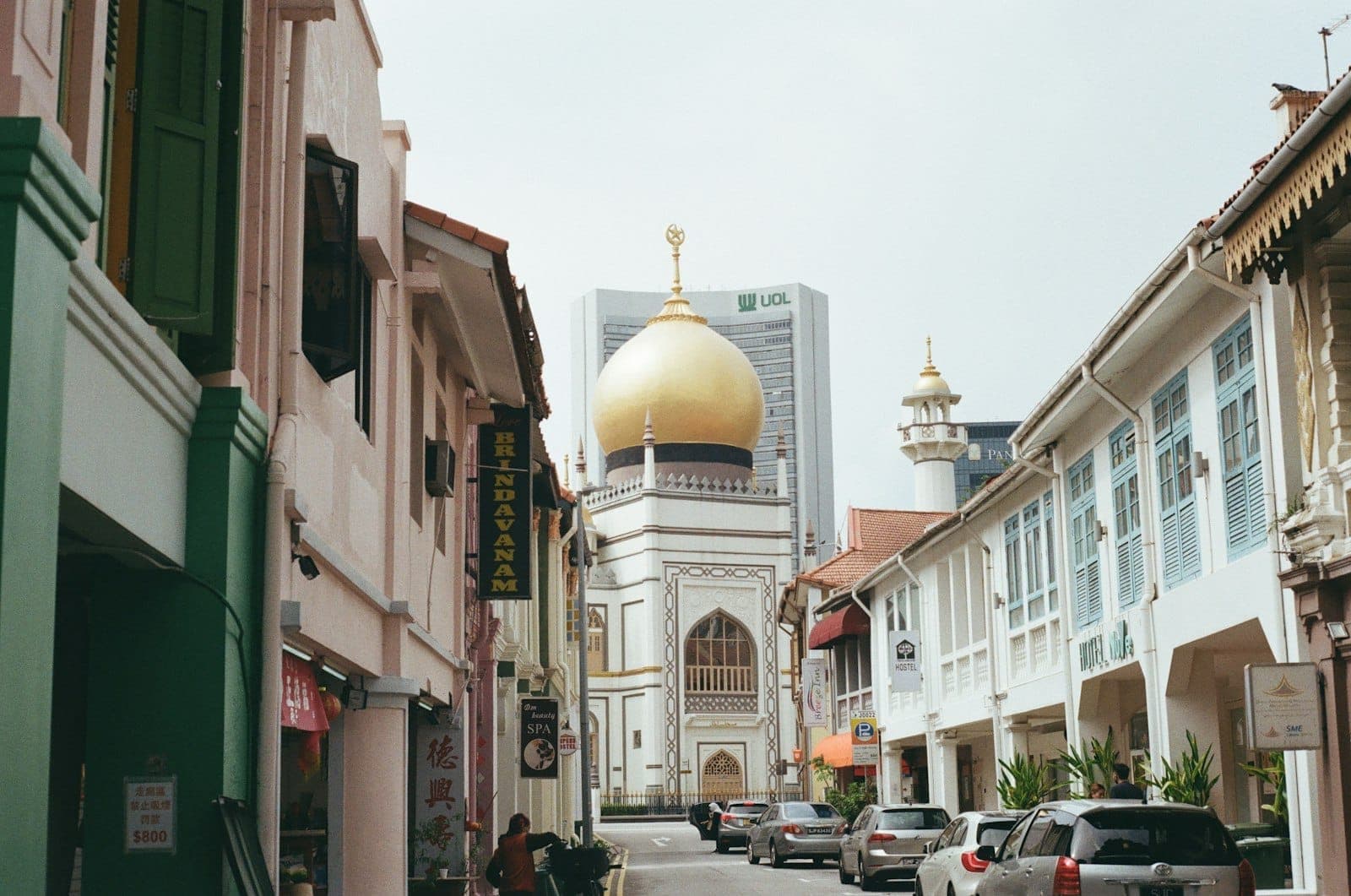 Street view of Kampong Glam with the golden dome of Sultan Mosque framed by colorful shophouses, cars parked along the road, and city buildings in the background.