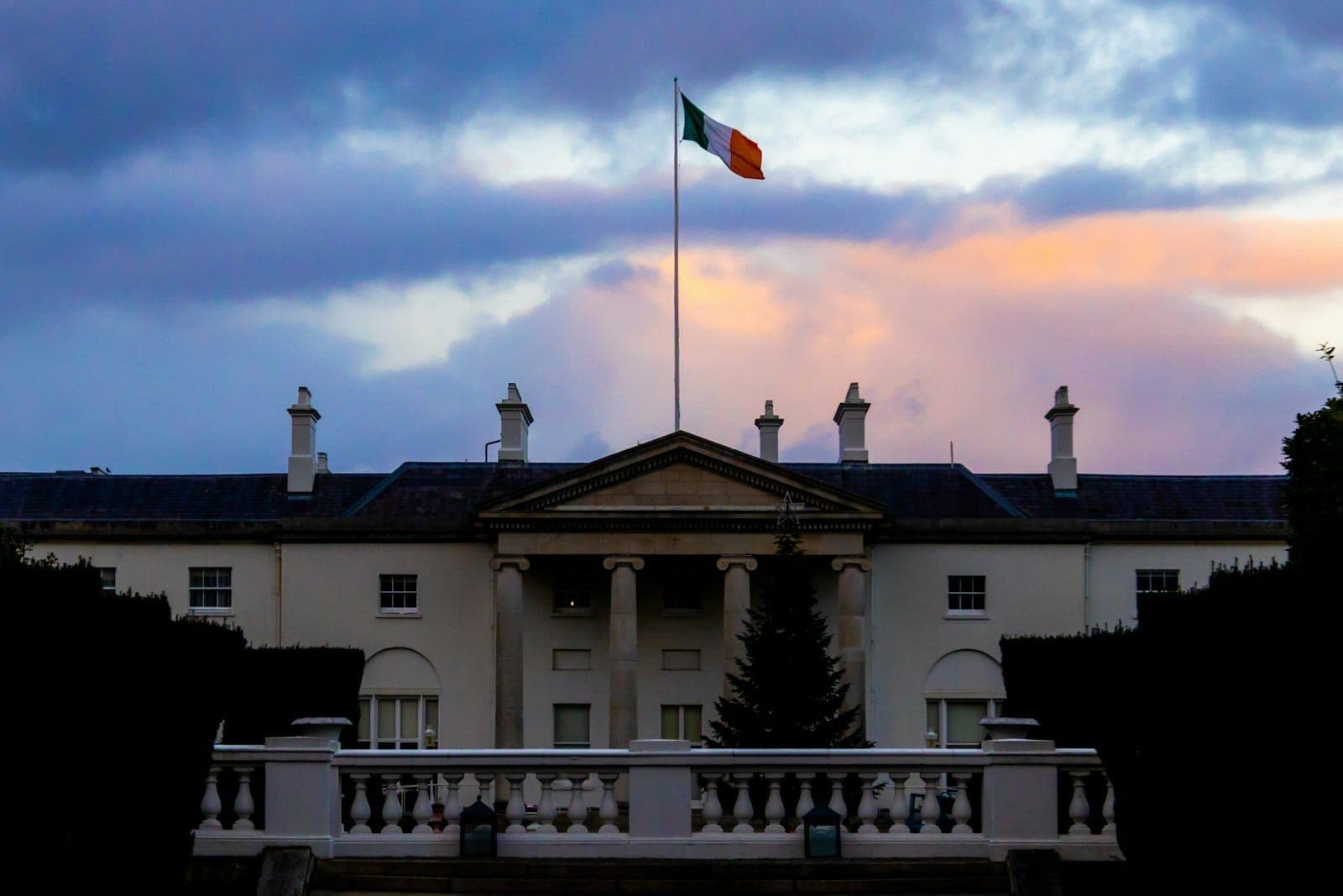 Vista frontal de Áras an Uachtaráin, residencia presidencial irlandesa, con la bandera irlandesa ondeando, pilares blancos y un dramático cielo nocturno.