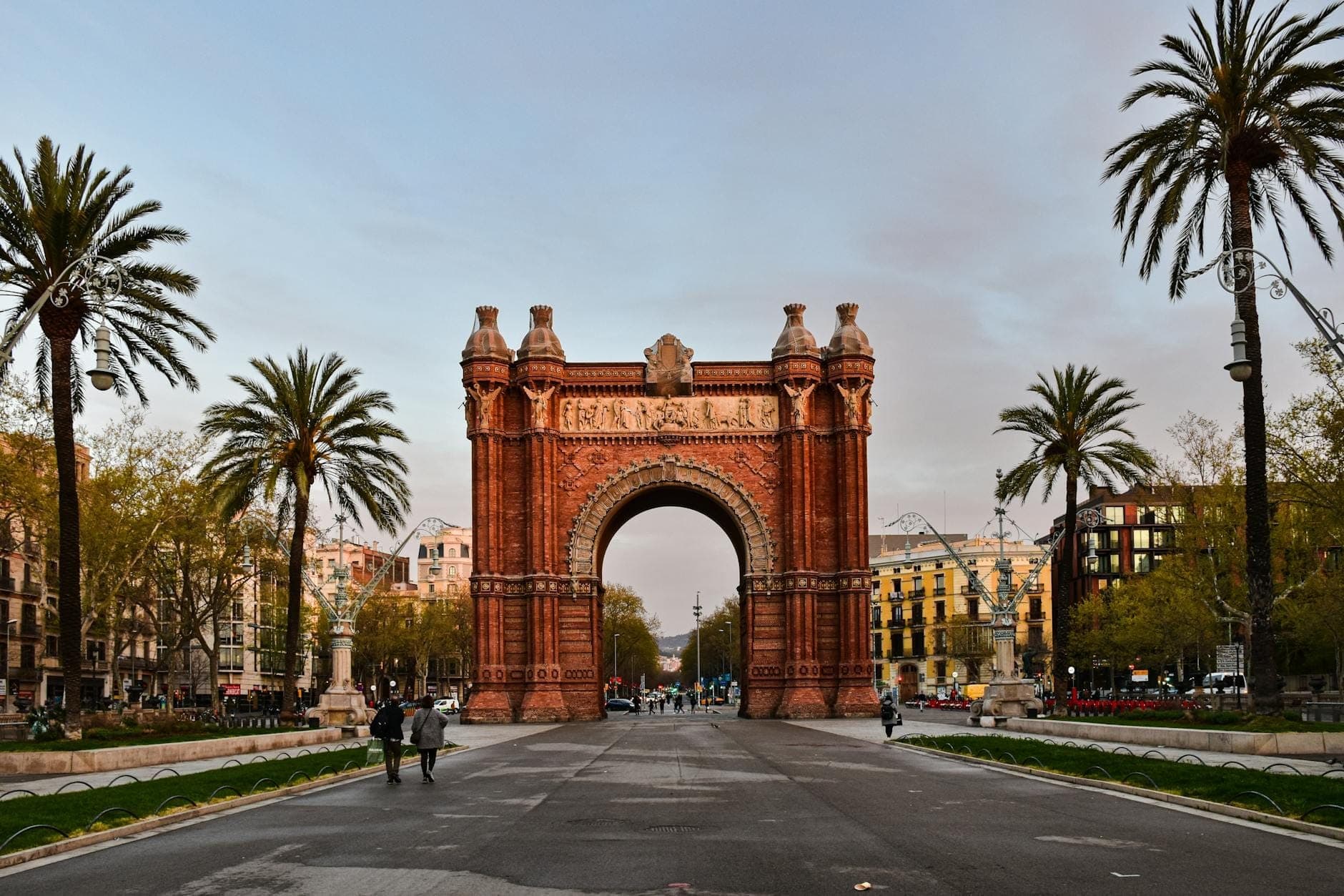 Toàn cảnh Arc de Triomf ở Barcelona với hàng cọ dọc hai bên đại lộ trung tâm. Mọi người đang đi bộ về phía và xung quanh công trình dưới bầu trời vàng óng mềm mại.