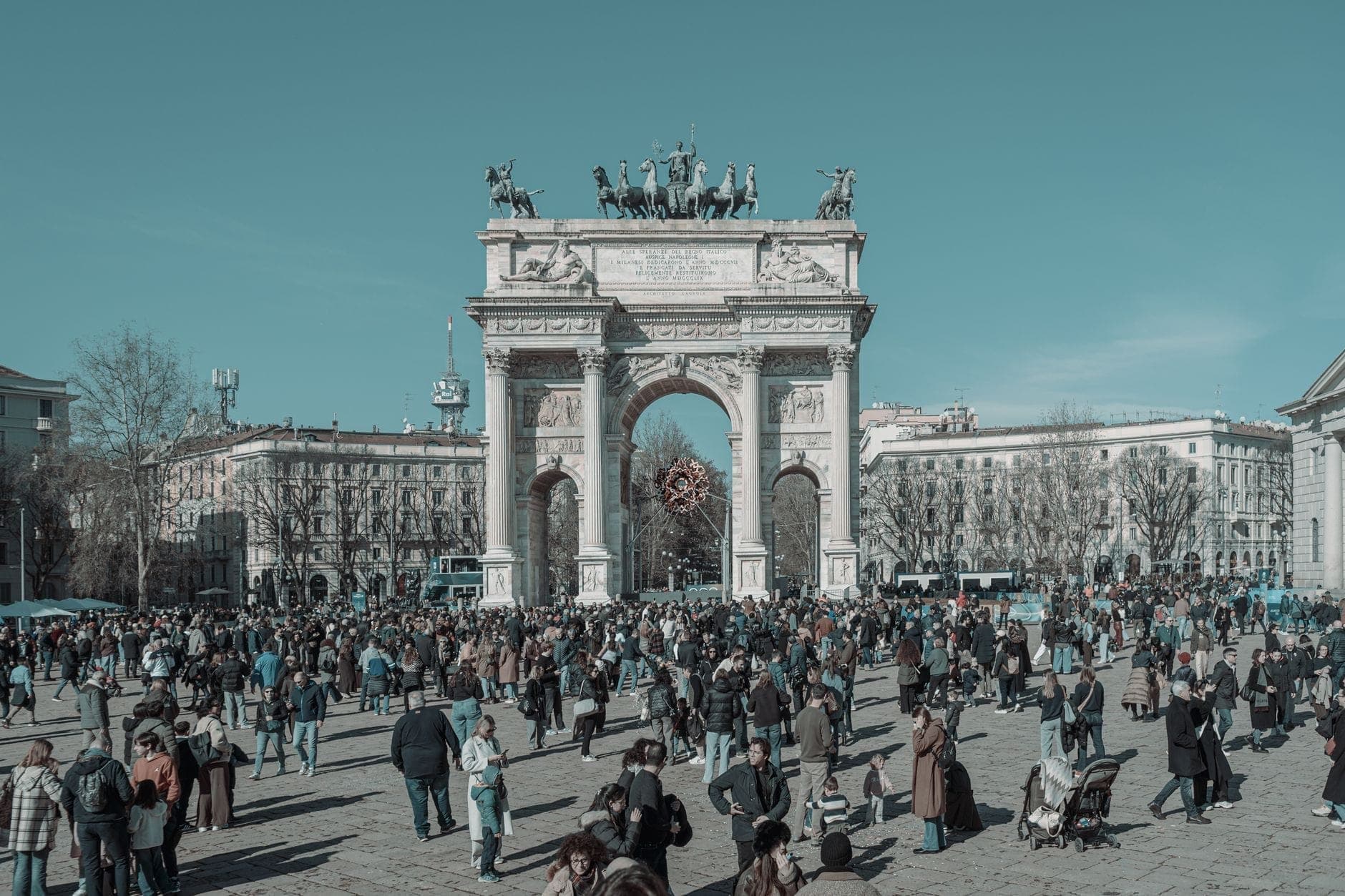 Crowd of people gathered in front of the Arco della Pace in Milan on a clear day, showcasing the full arch and lively square.