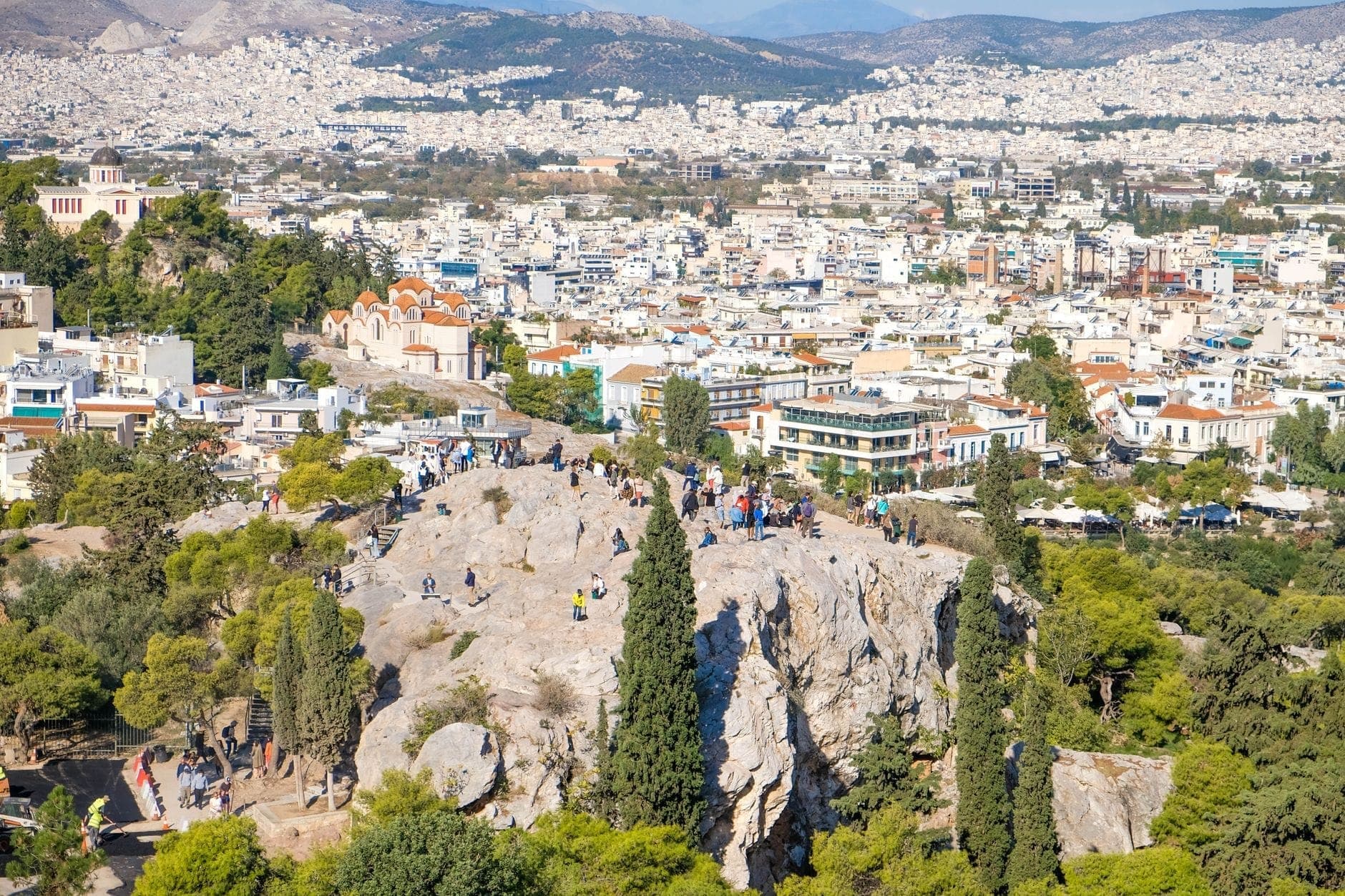 A sunlit view of Areopagus Hill with people on the rocky outcrop, surrounded by trees and Athens cityscape in the background.