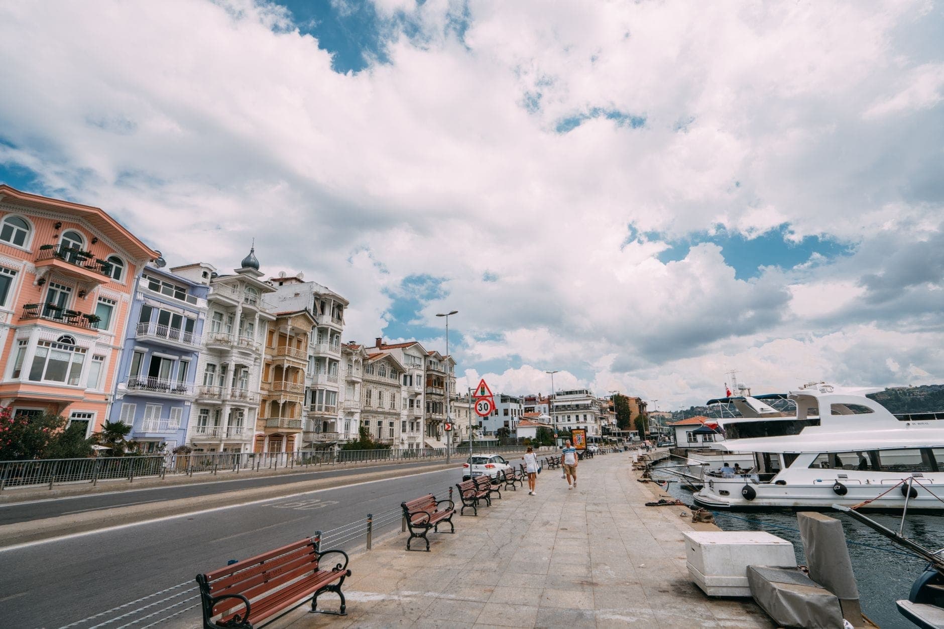 Die Uferpromenade von Arnavutköy mit farbenfrohen historischen osmanischen Holzhäusern, Bänken entlang des Bosporus und am Wasser vertäuten Booten unter strahlend blauem Himmel.