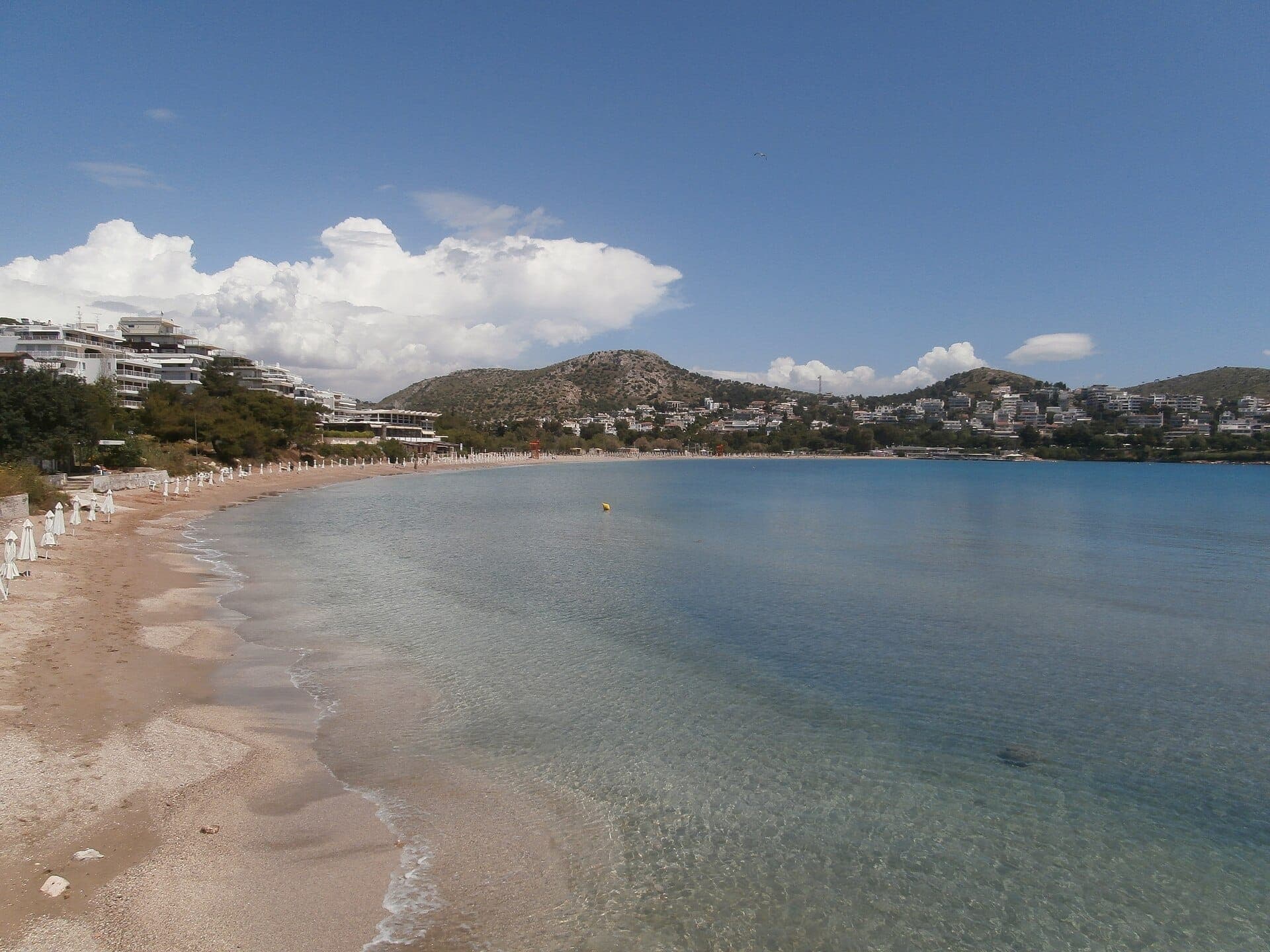 Wide sandy beach on the Athenian Riviera with clear blue water, white umbrellas, modern buildings, and green hills under a bright sky.