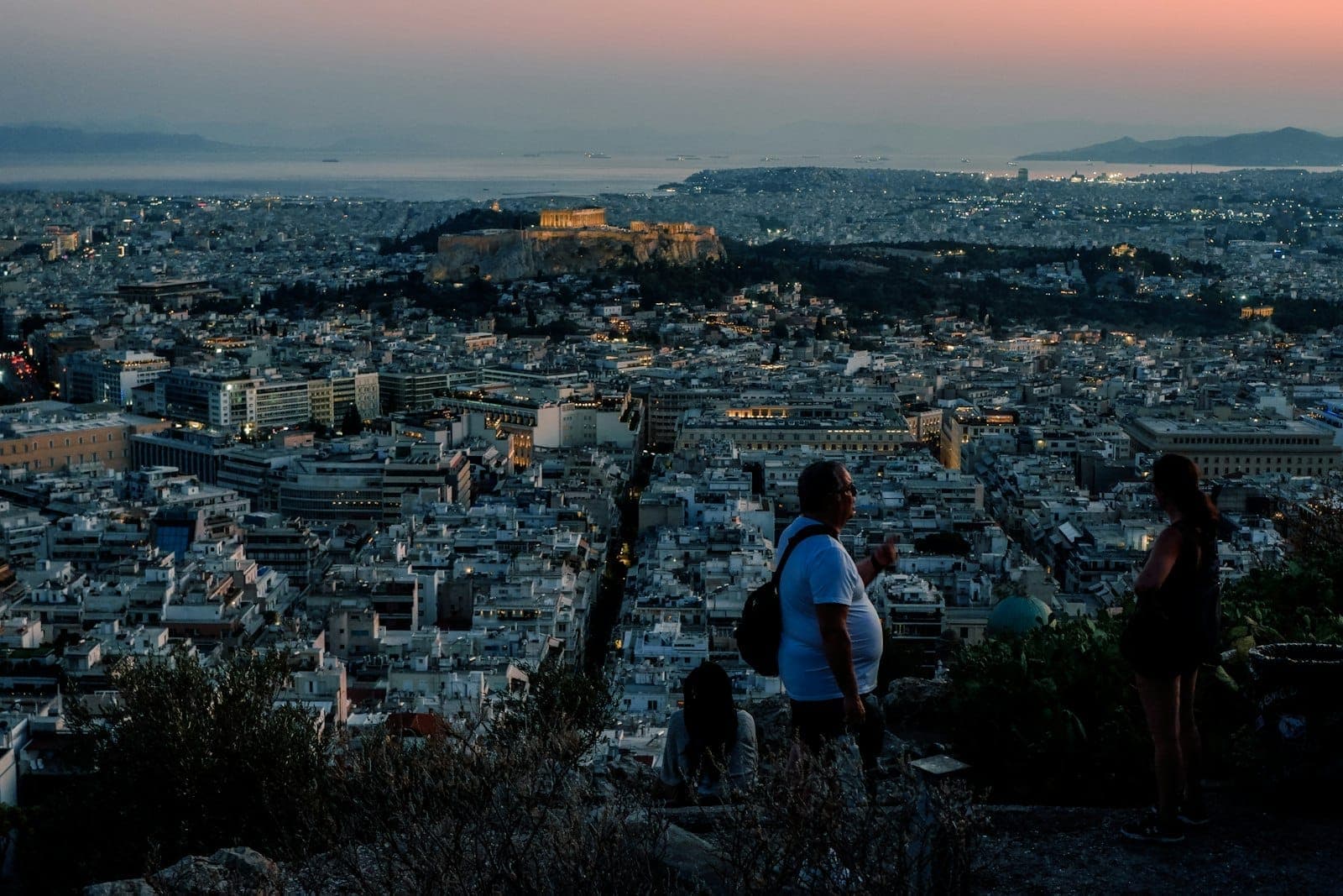 Blick von einem Hügel in Athen bei Sonnenuntergang mit Menschen im Vordergrund, die über die Stadt und die beleuchtete Akropolis in der Ferne schauen.