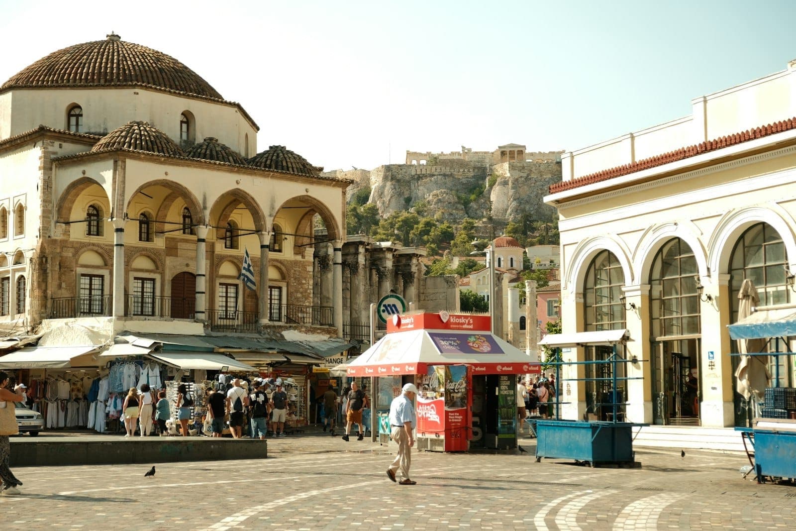 Blick auf den Monastiraki-Platz in Athen mit der Akropolis im Hintergrund und Menschen an den Ständen des Freiluftmarkts.