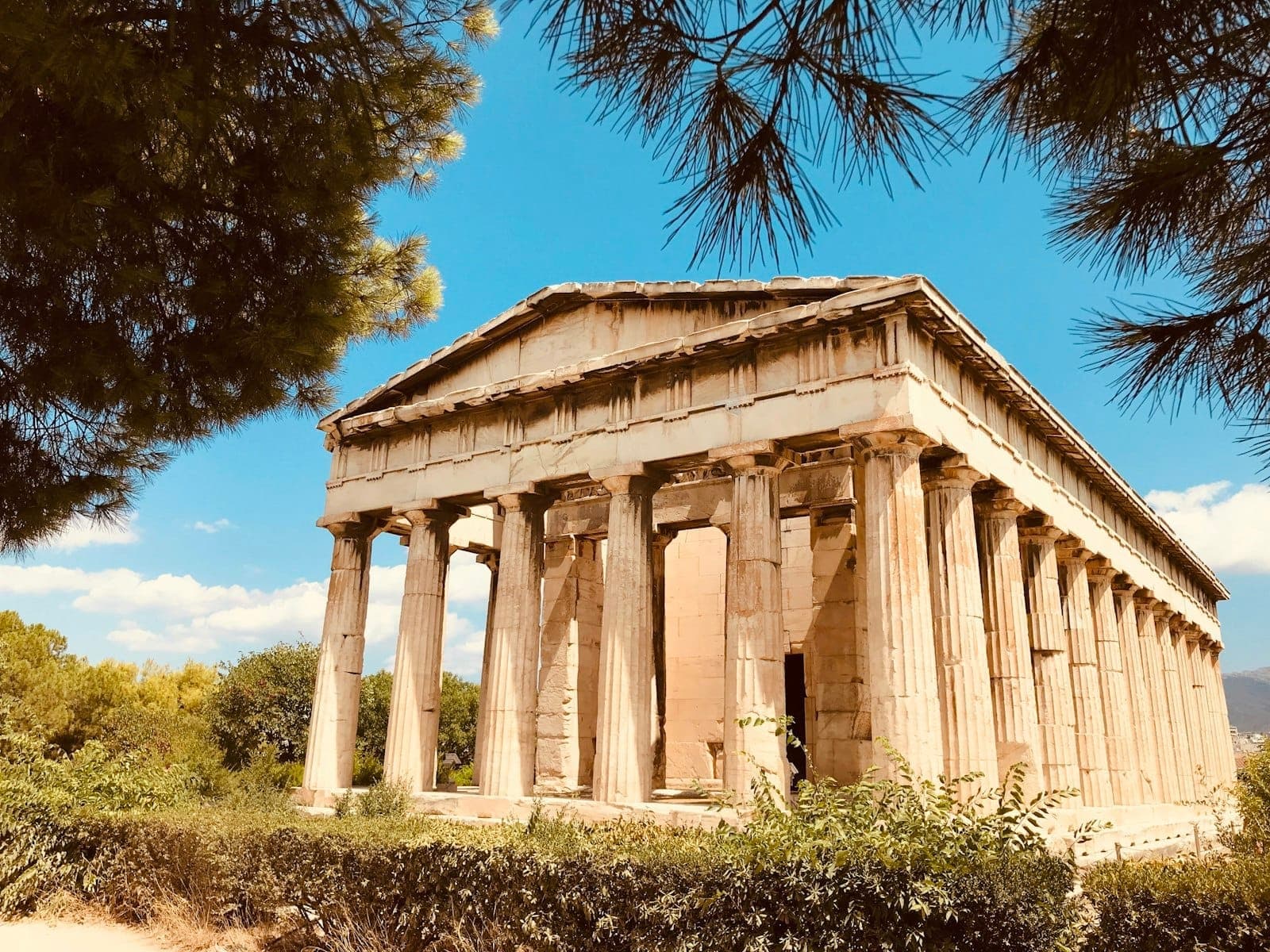 Kuil Hephaestus di Athena, dikelilingi pepohonan hijau di bawah langit biru cerah, dilihat dari sudut yang memperlihatkan kolom-kolom dan pedimen.