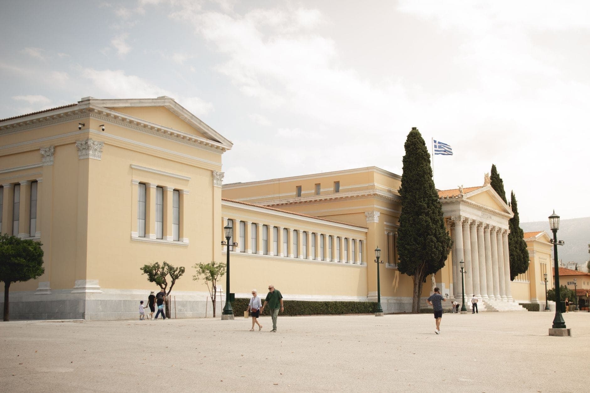 Pemandangan luas gedung neoklasik Museum Arkeologi Nasional di Athena dengan orang-orang berjalan di luar.