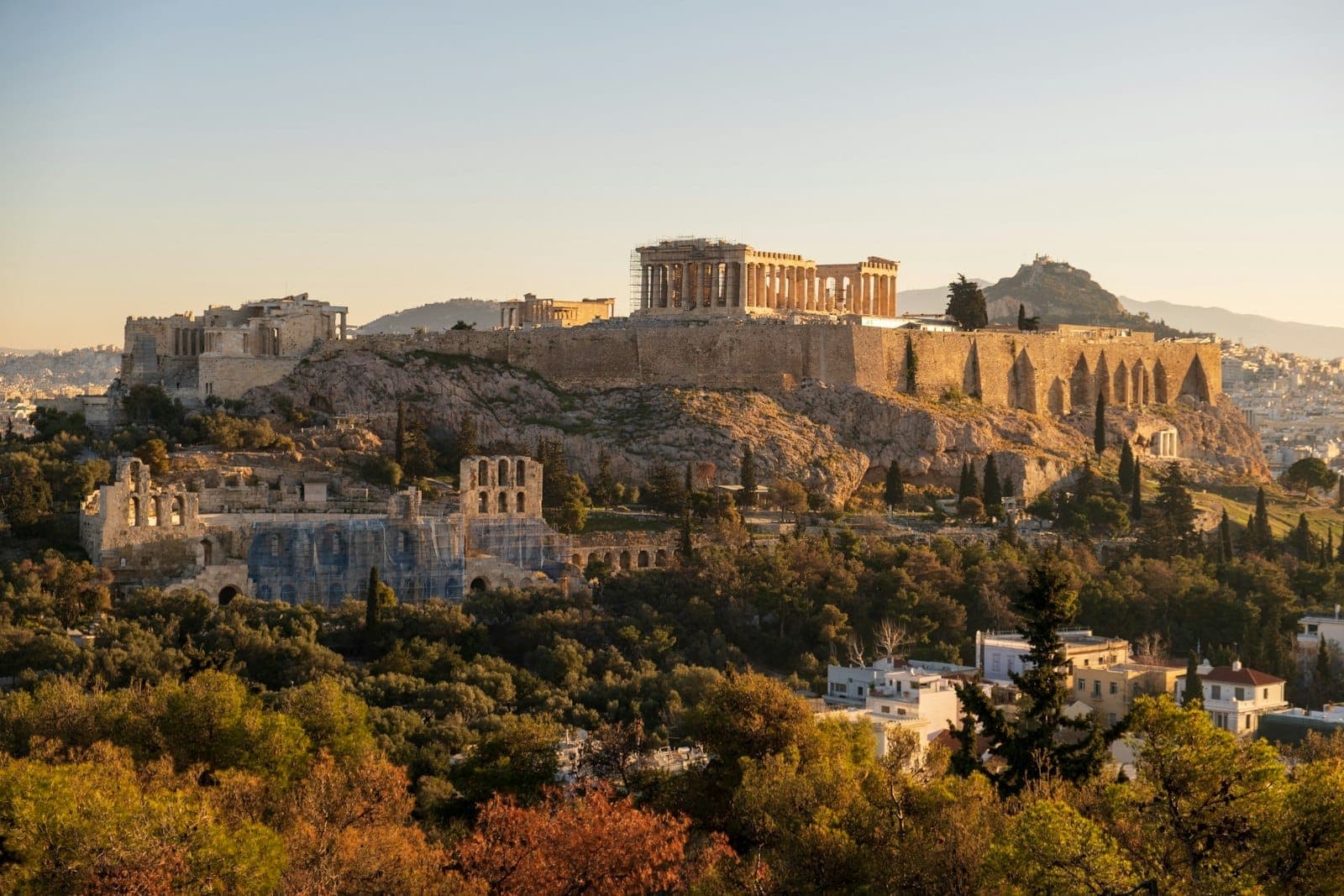 Pemandangan luas dan terang bukit Acropolis dengan Parthenon dan monumen kuno di sekitarnya di Athena, dibingkai oleh pepohonan hijau saat matahari terbenam.