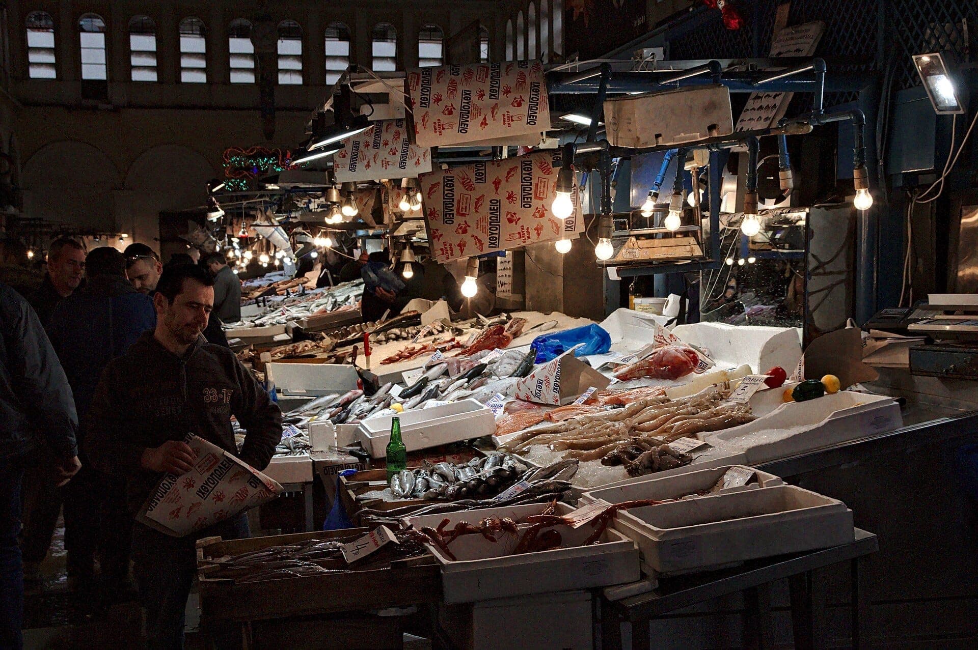 Fishmongers and shoppers gather around illuminated seafood stalls at Athens Central Market, with fresh fish and octopus displayed on ice under hanging bulbs.