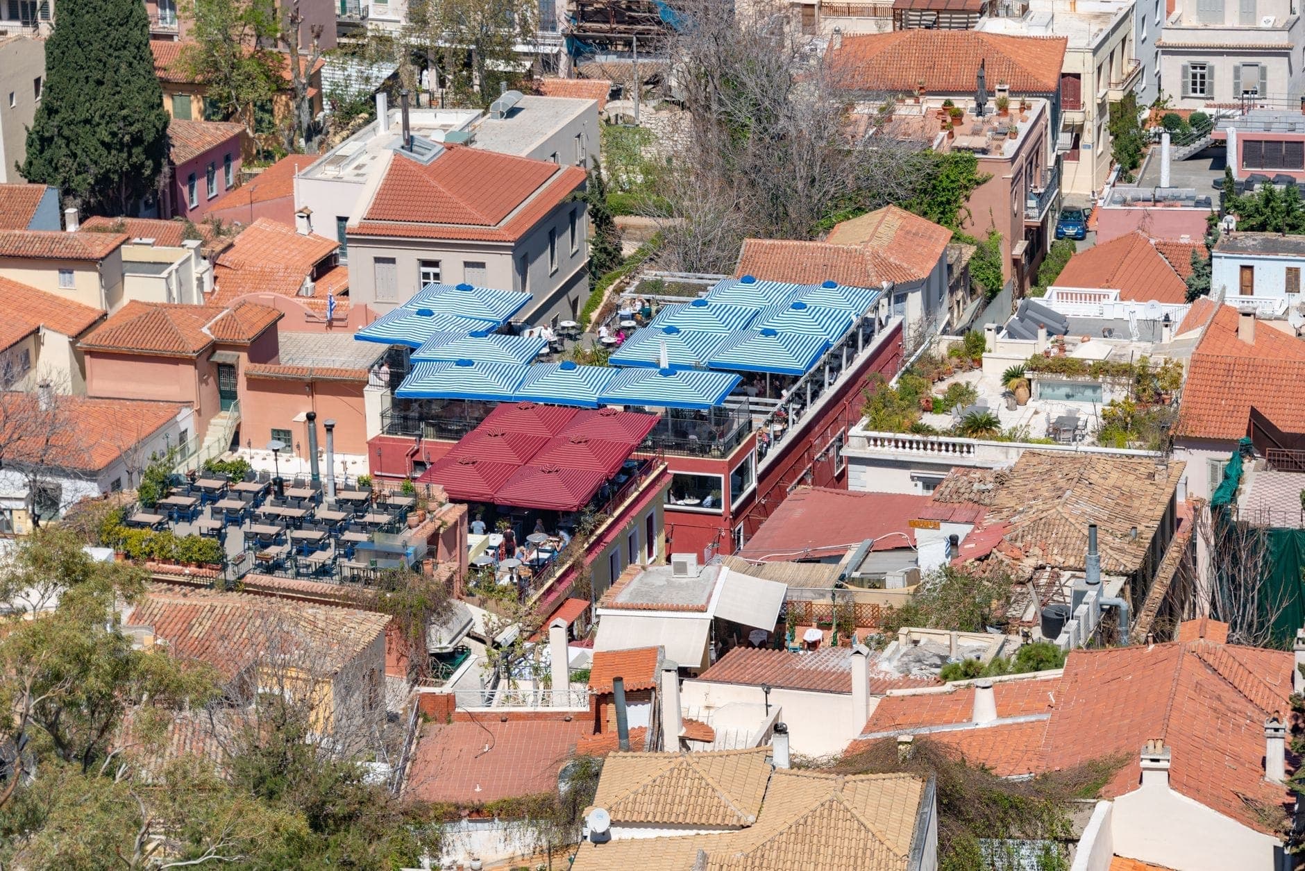 Luftaufnahme eines Stadtviertels in Athen mit gruppierten Restaurants mit blauen und roten Markisen zwischen traditionellen Ziegeldächern.