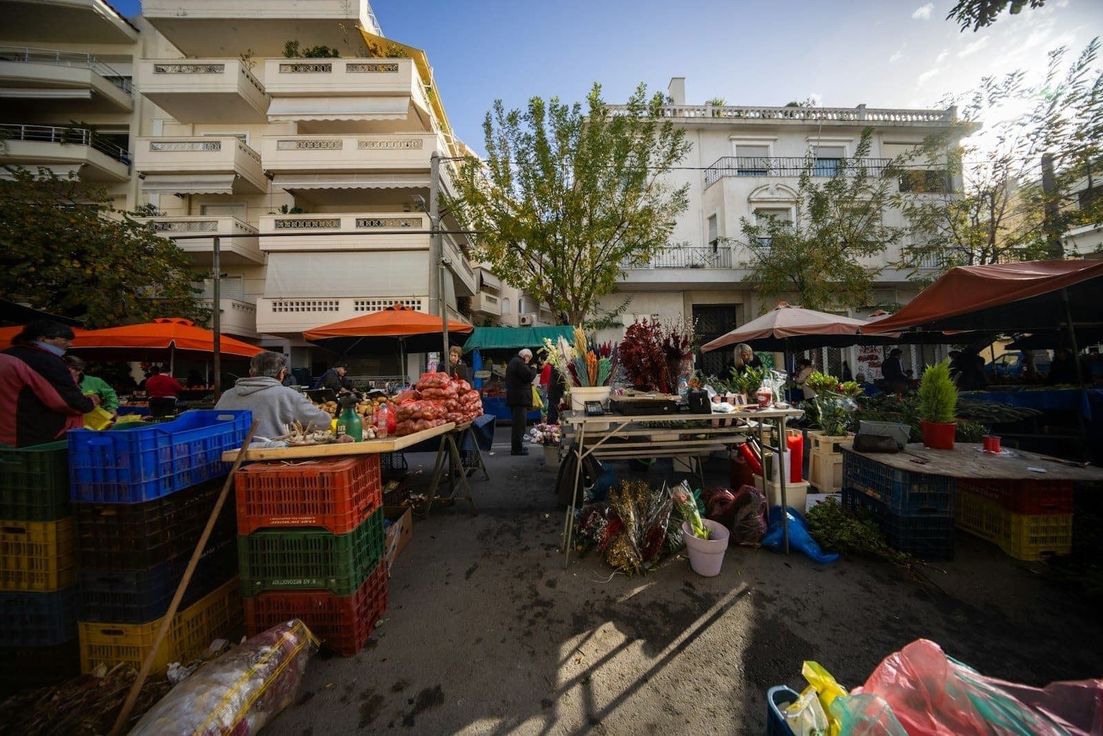 Markt im Freien in Athen mit frischen Früchten und Gemüse auf Tischen unter orangefarbenen Zelten, einkaufenden Menschen und Wohngebäuden im Hintergrund.