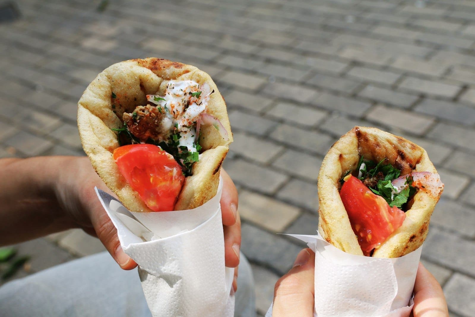 Two hands holding fresh Greek gyros filled with meat, tomato, herbs, and sauce, outdoors on a sunlit stone street in Athens.