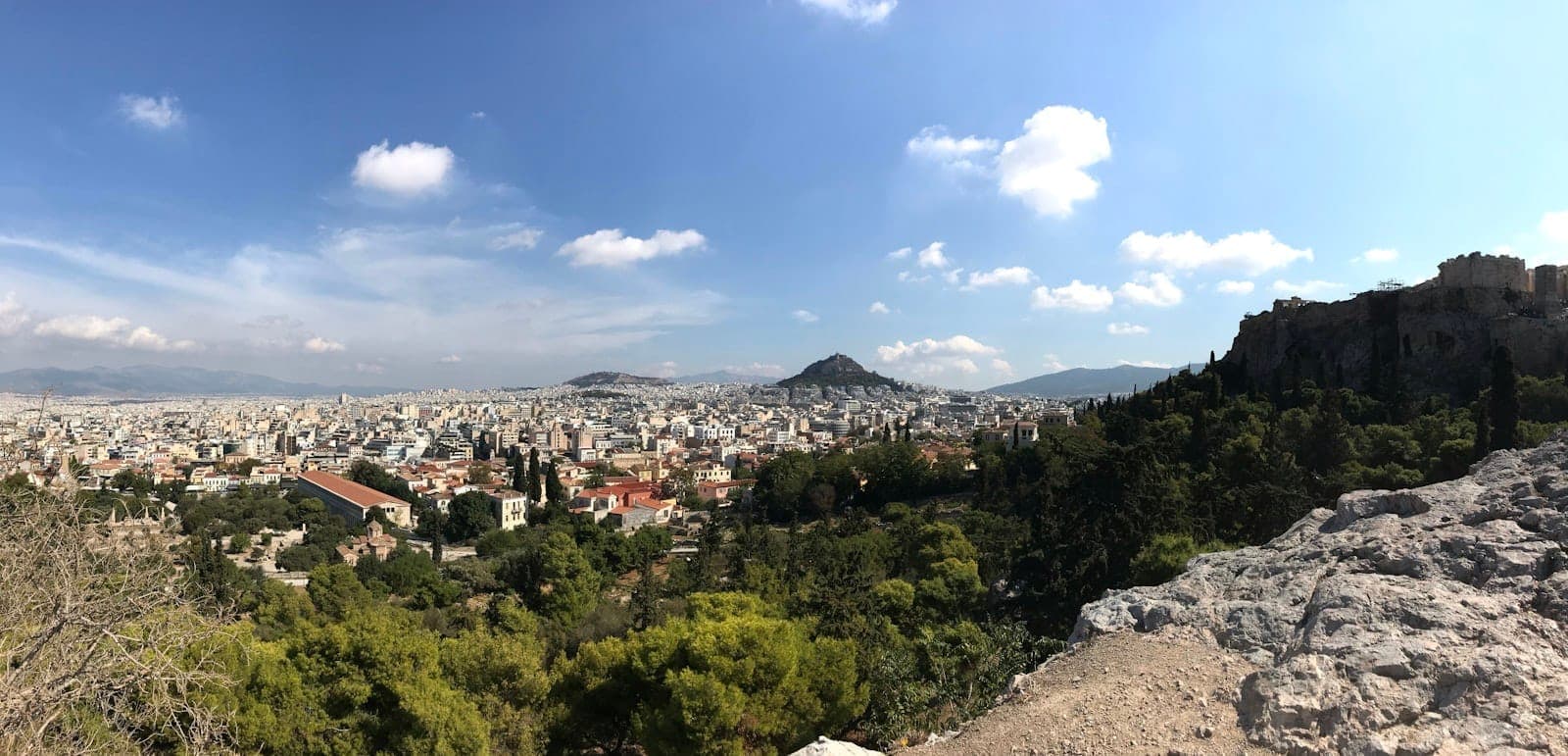 Panoramic view of Athens from a hilltop, showing city buildings, green trees, distant hills, and blue sky with scattered clouds. A rocky outcrop frames the foreground.