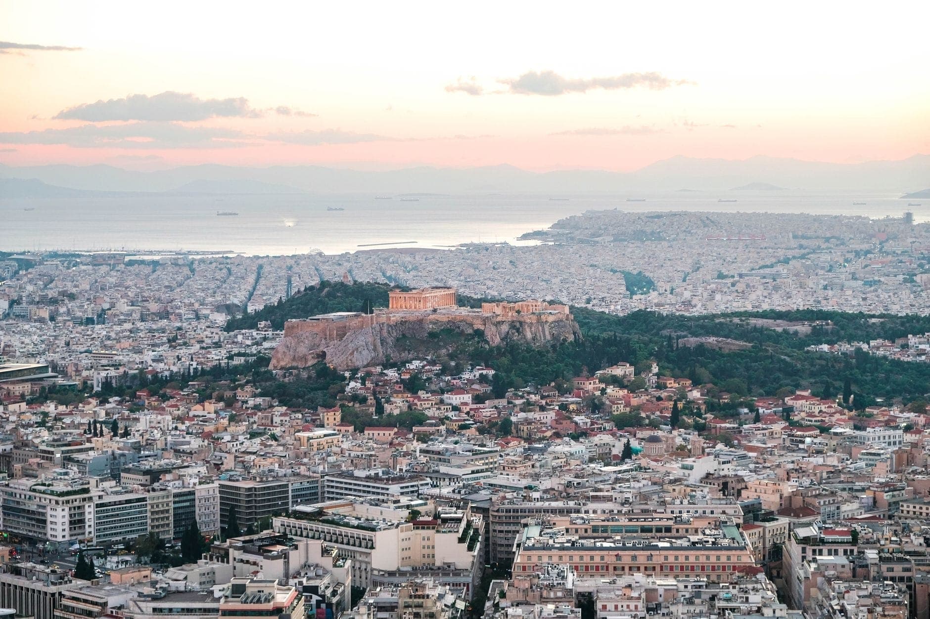 A wide panoramic view of Athens at sunset with the Acropolis illuminated on a hill, sprawling cityscape, and subtle pink sky above the sea in the distance.