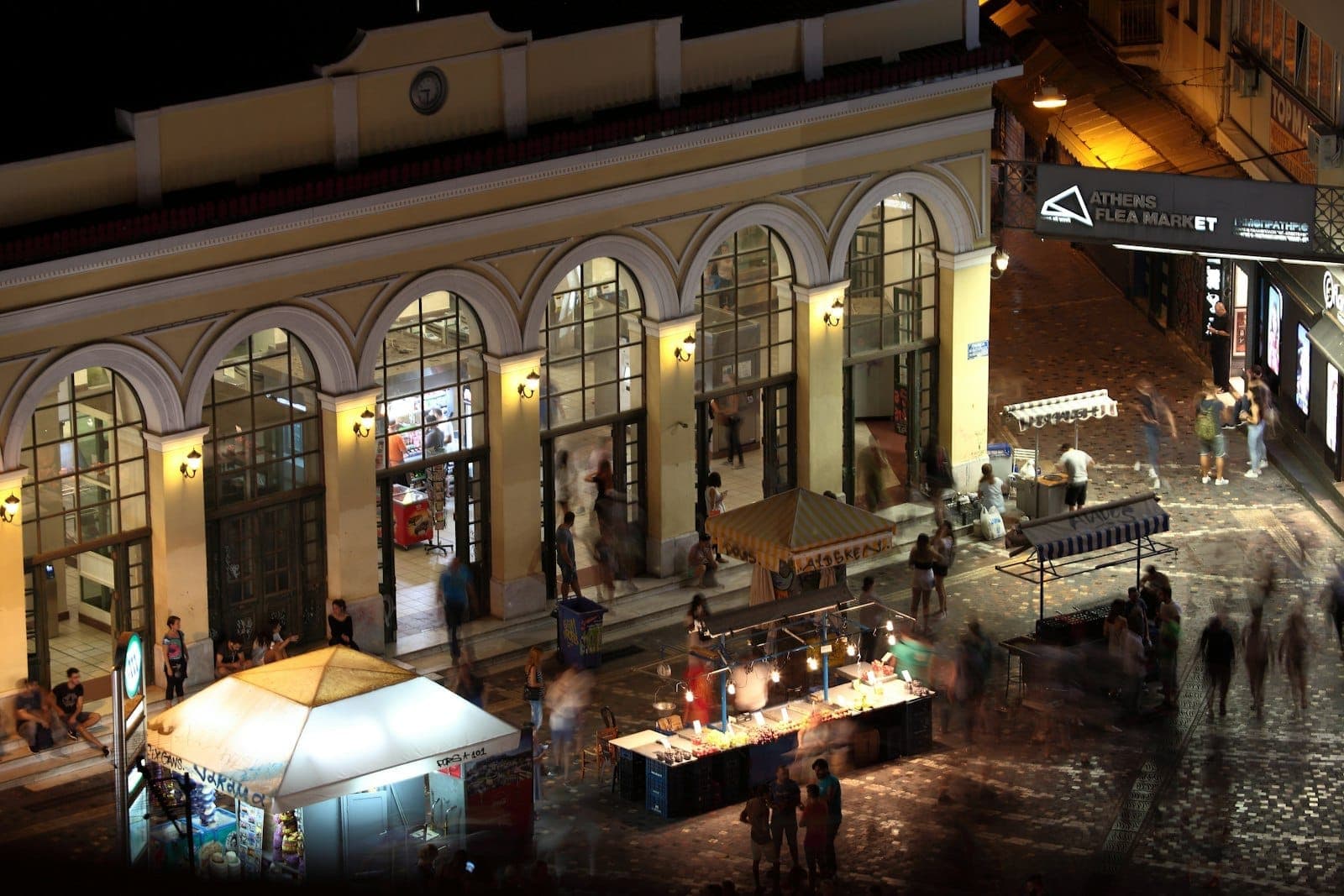 A lively nighttime scene in Athens with people gathering outside a brightly lit building, market stalls, and illuminated streets, capturing the city’s energetic evening atmosphere.
