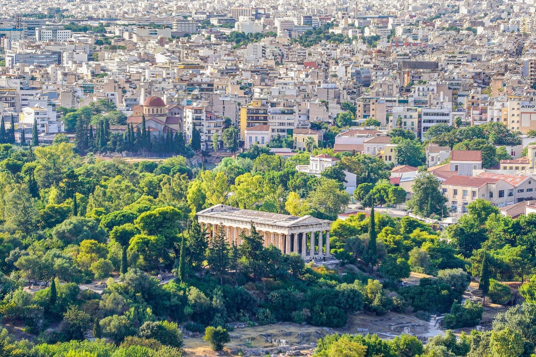 Luftaufnahme von Athen mit dem antiken Tempel des Hephaistos, umgeben von grünen Bäumen und der Stadtsilhouette im Hintergrund.