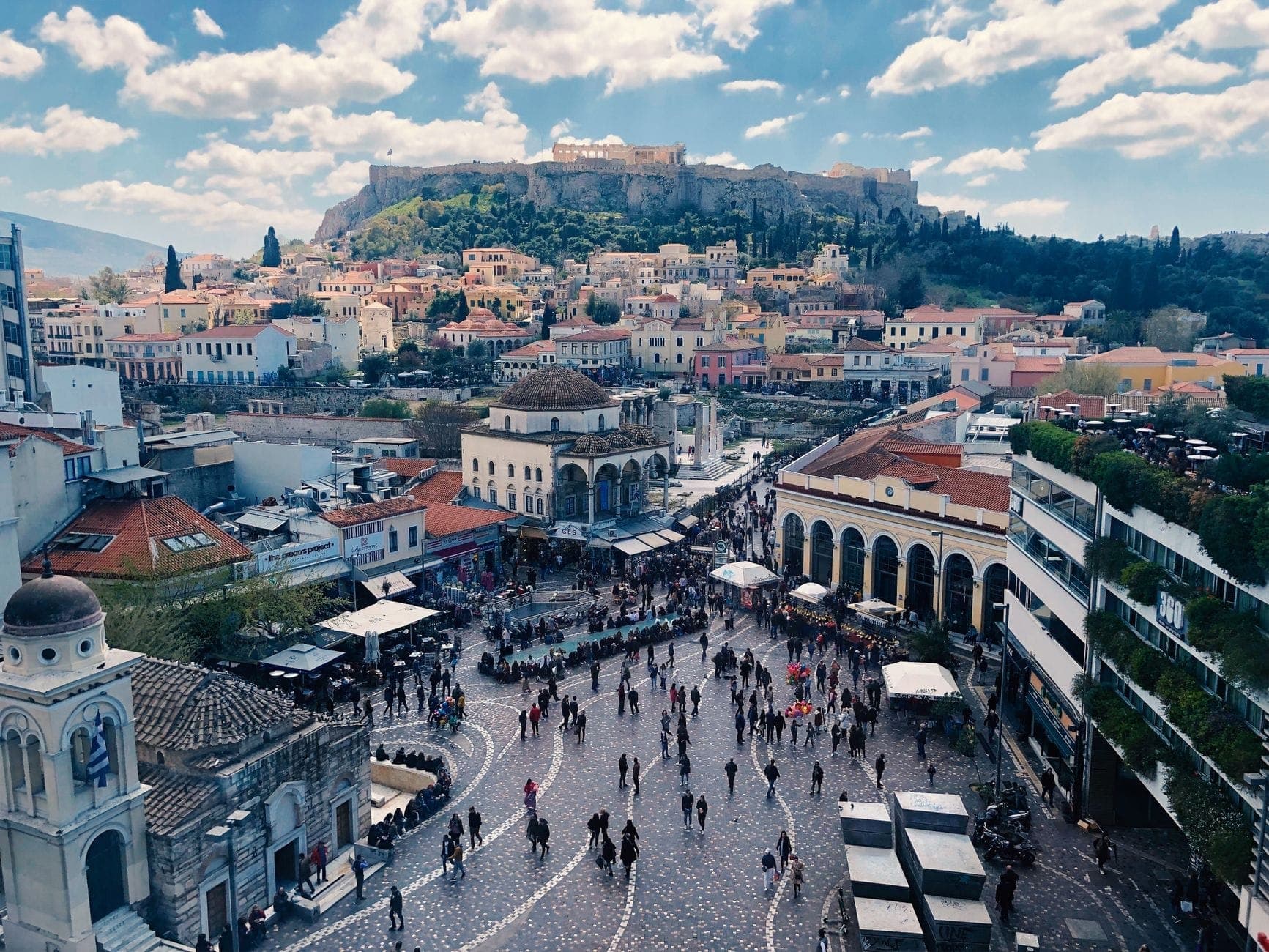 Luftaufnahme des Monastiraki-Platzes in Athen mit der Akropolis im Hintergrund, belebt von Menschen und umgeben von budgetfreundlichen Gebäuden.