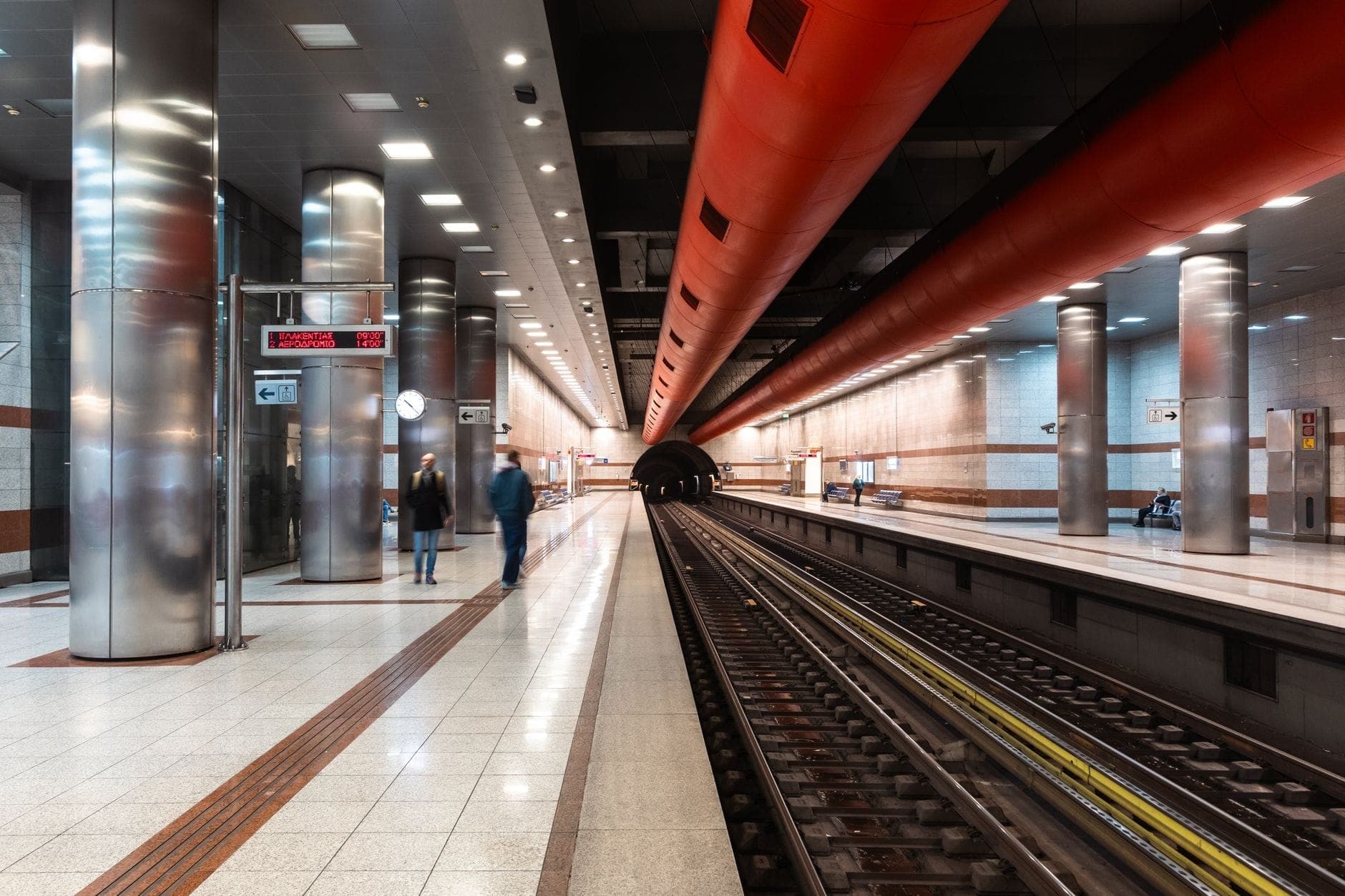 Moderne U-Bahn-Station Athens mit Gleisen, roter Deckengestaltung und einigen wartenden Personen auf dem Bahnsteig.