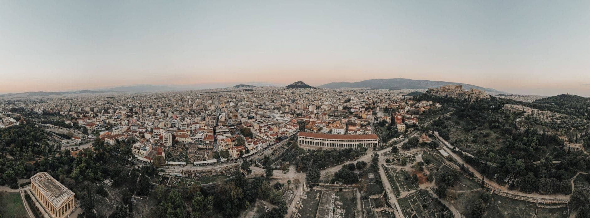 Wide panoramic aerial view of Athens at sunrise with the Acropolis and surrounding neighborhoods, showcasing the city’s scale and landscape under a soft, calm sky.