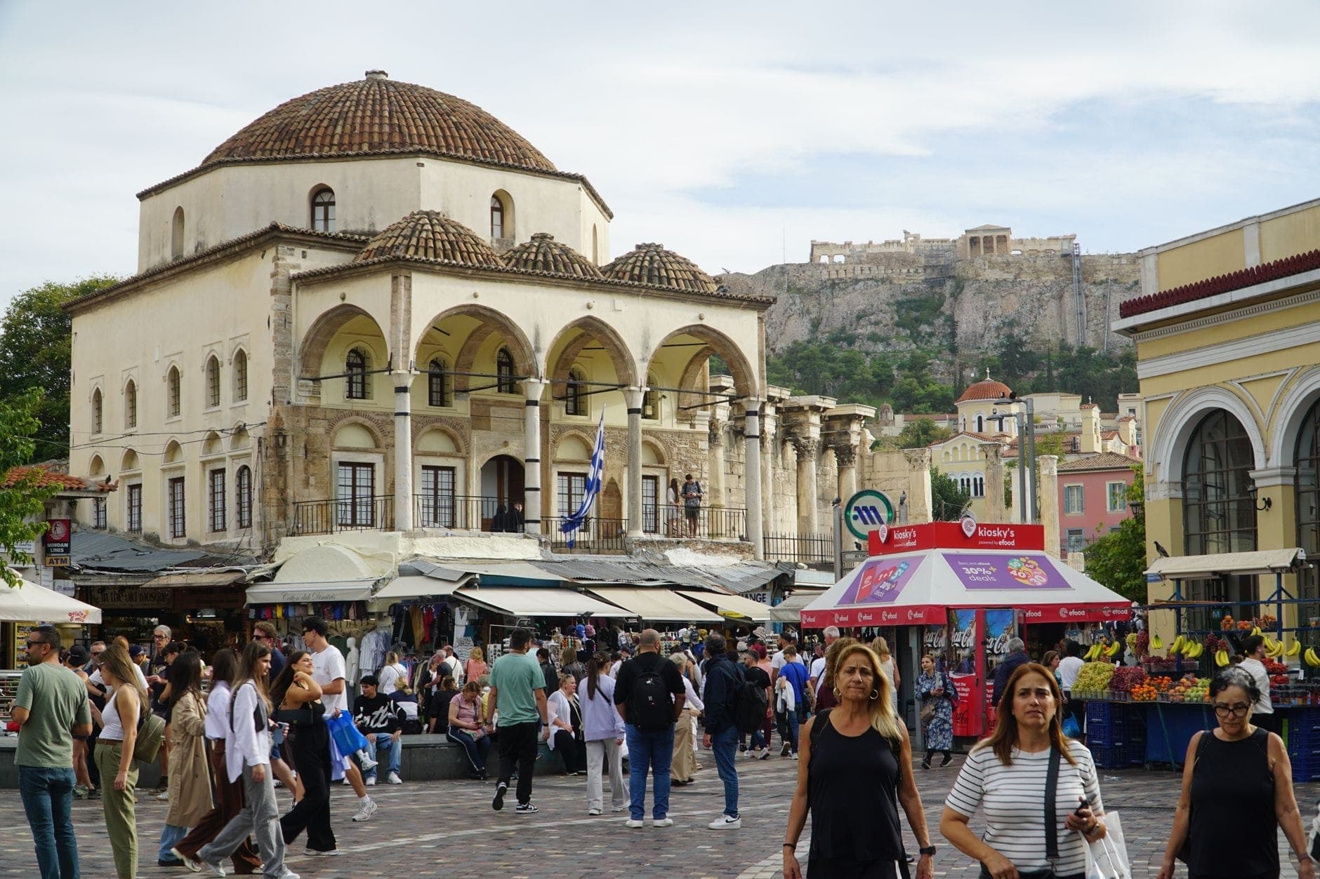 Busy Monastiraki Square in Athens with vendors, market stalls, and shoppers, set against the backdrop of historic buildings and the Acropolis above.
