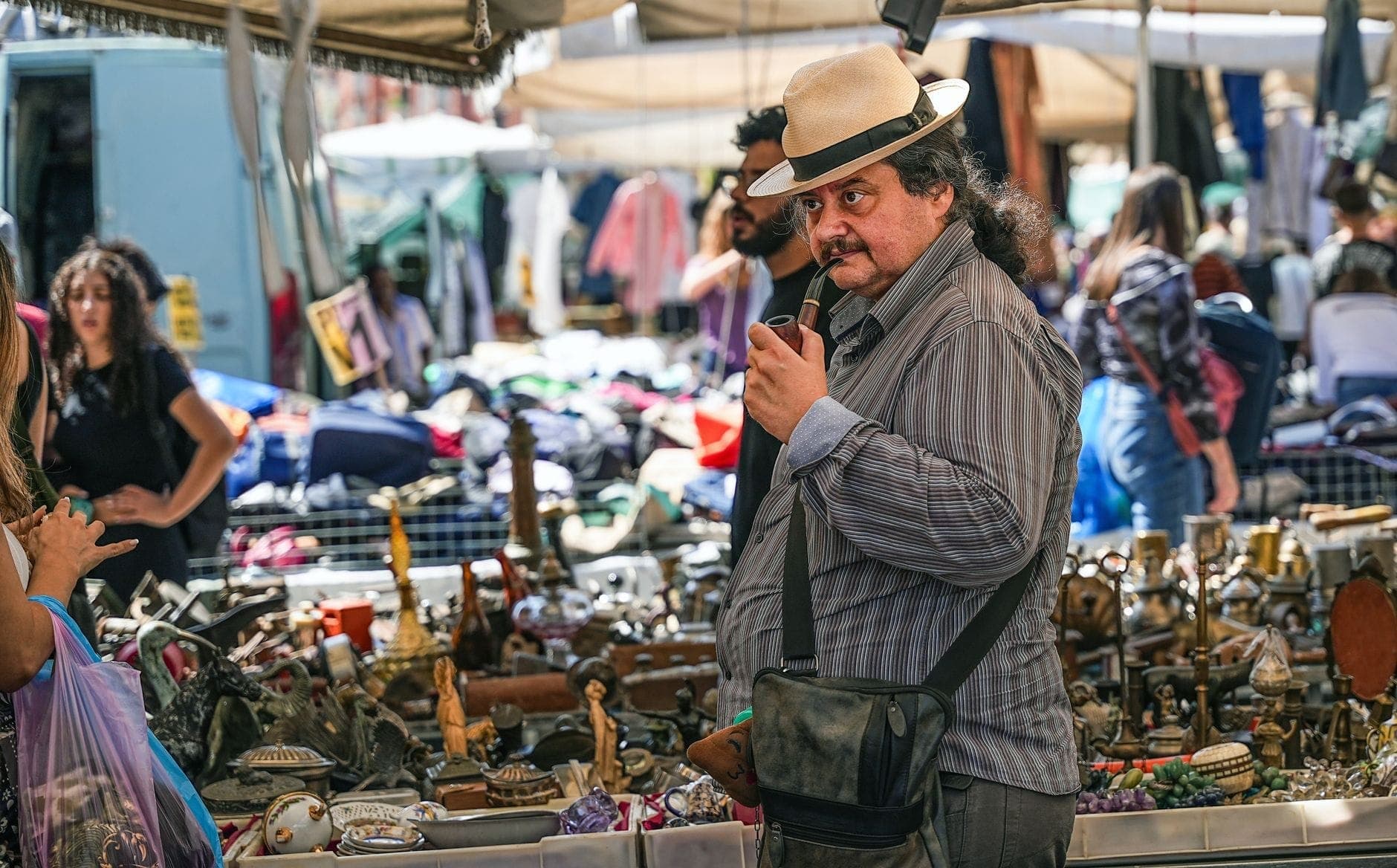 Hombre con sombrero examinando pequeñas antigüedades en un puesto de mercadillo al aire libre repleto de diversos artículos de segunda mano y personas curioseando.