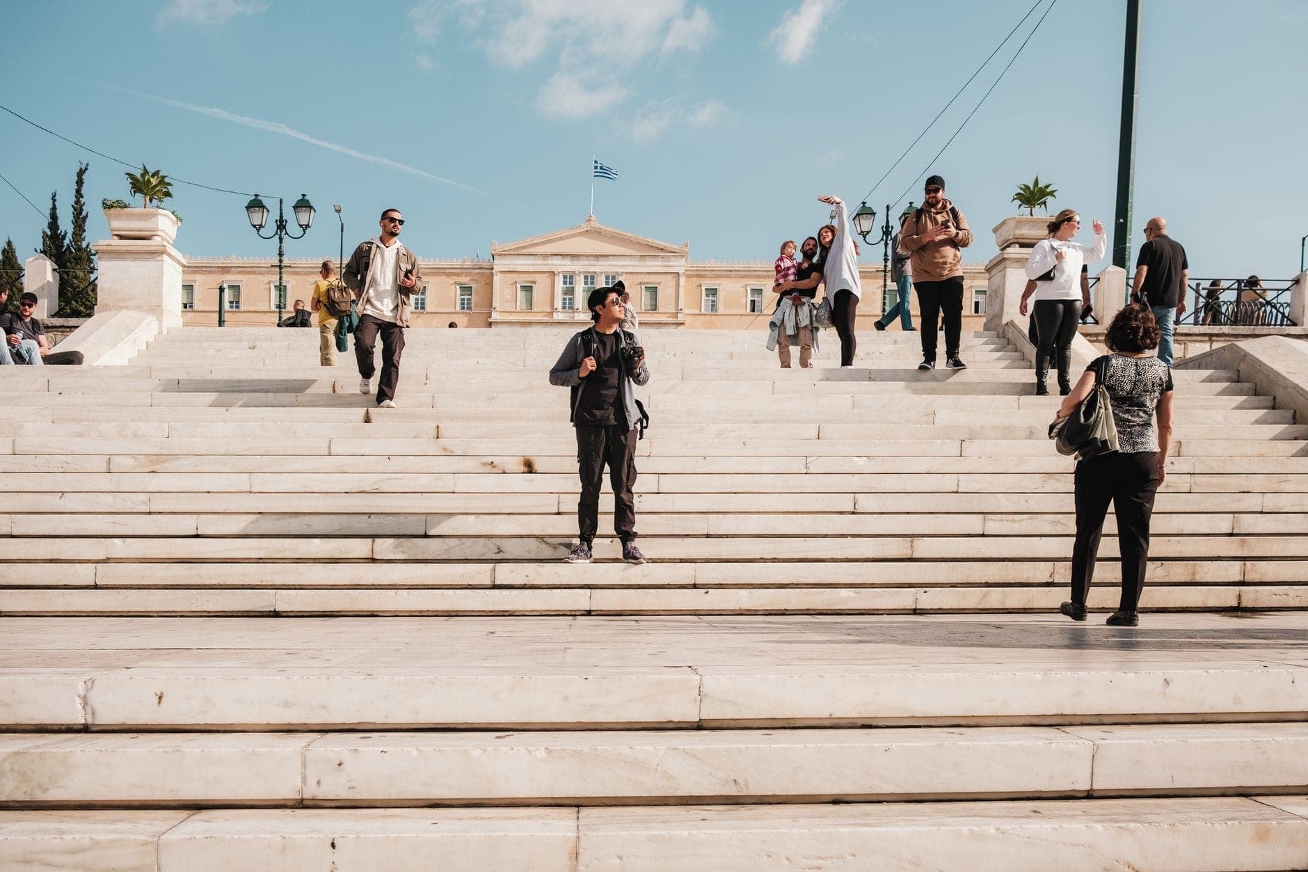 Touristen spazieren und fotografieren auf den Marmorstufen des Syntagma Square mit dem Gebäude des Hellenischen Parlaments im Hintergrund unter einem blauen Himmel.