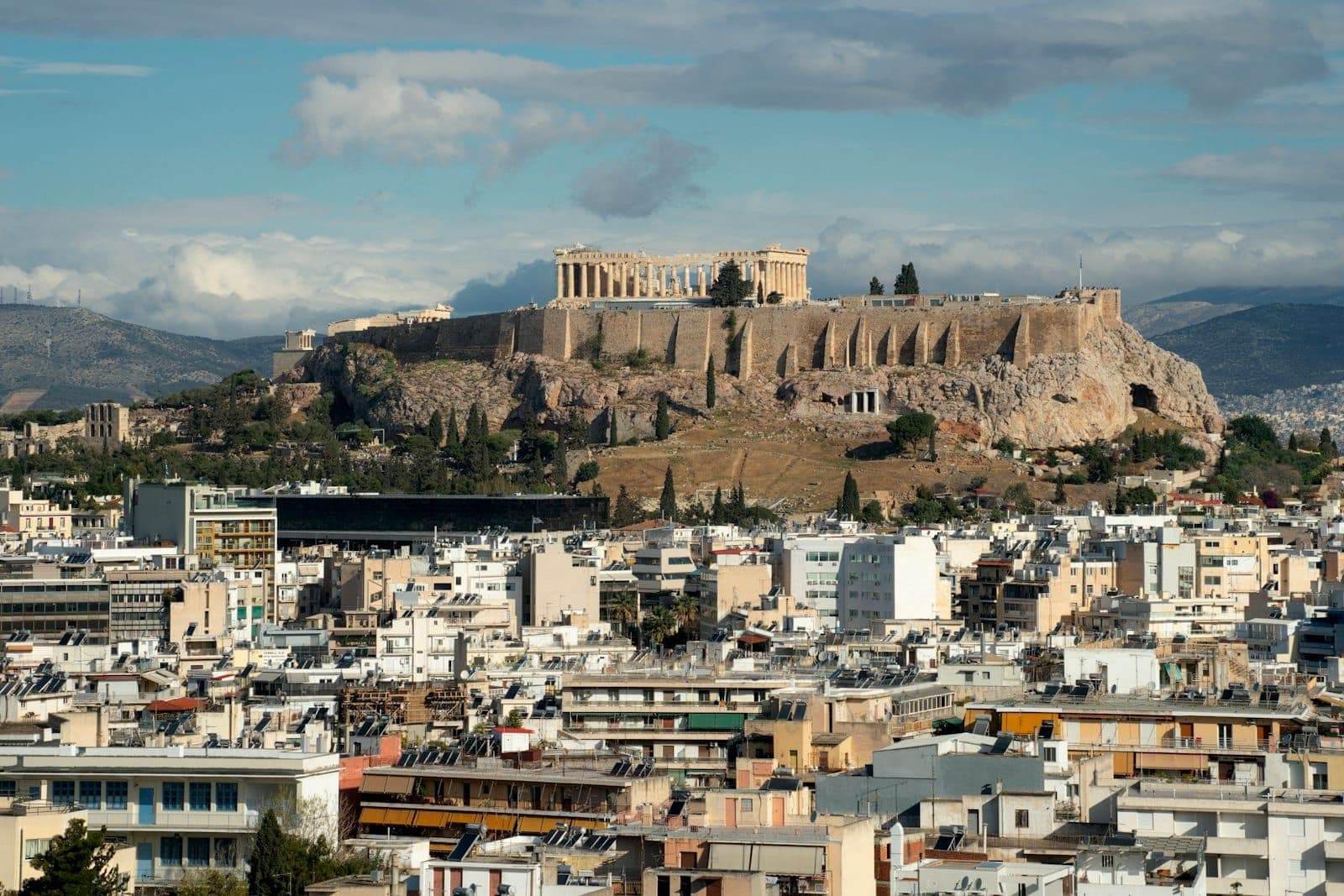 Wide view of the Athens cityscape with the iconic Acropolis and Parthenon prominently visible above surrounding modern buildings under a bright sky.