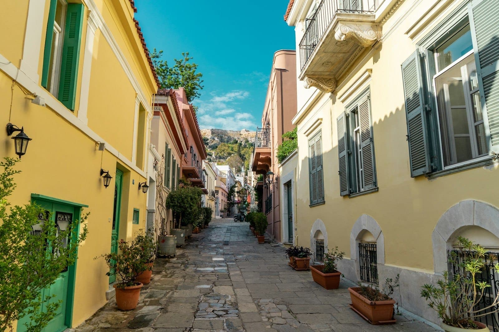 Incantevole strada pedonale ad Atene con case colorate, piante in vaso e l'Acropoli visibile sullo sfondo sotto un cielo blu.