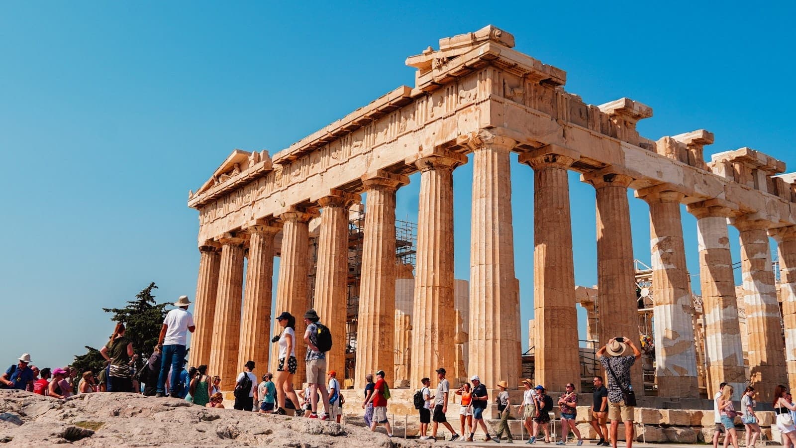 Families and children walking around the Parthenon temple on the Acropolis, under a bright blue sky in Athens, Greece.